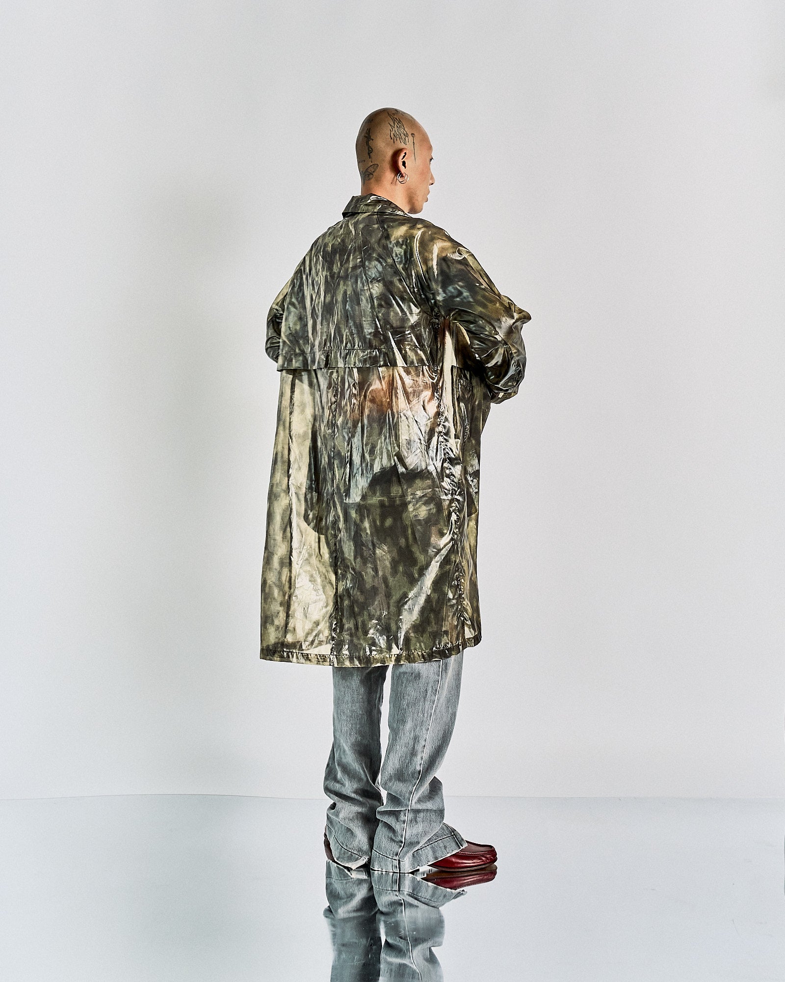 A person stands with their back to the camera, wearing a long, reflective, tie-dye Dries Van Noten coat and gray pants. The setting is minimal, emphasizing the outfit's texture.