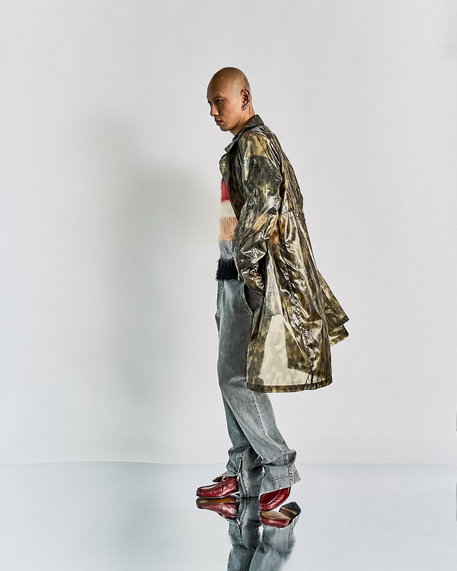 A person in a long, metallic Dries Van Noten coat stands sideways on a reflective surface. They wear gray jeans, a colorful sweater, and red boots, exuding a futuristic vibe.