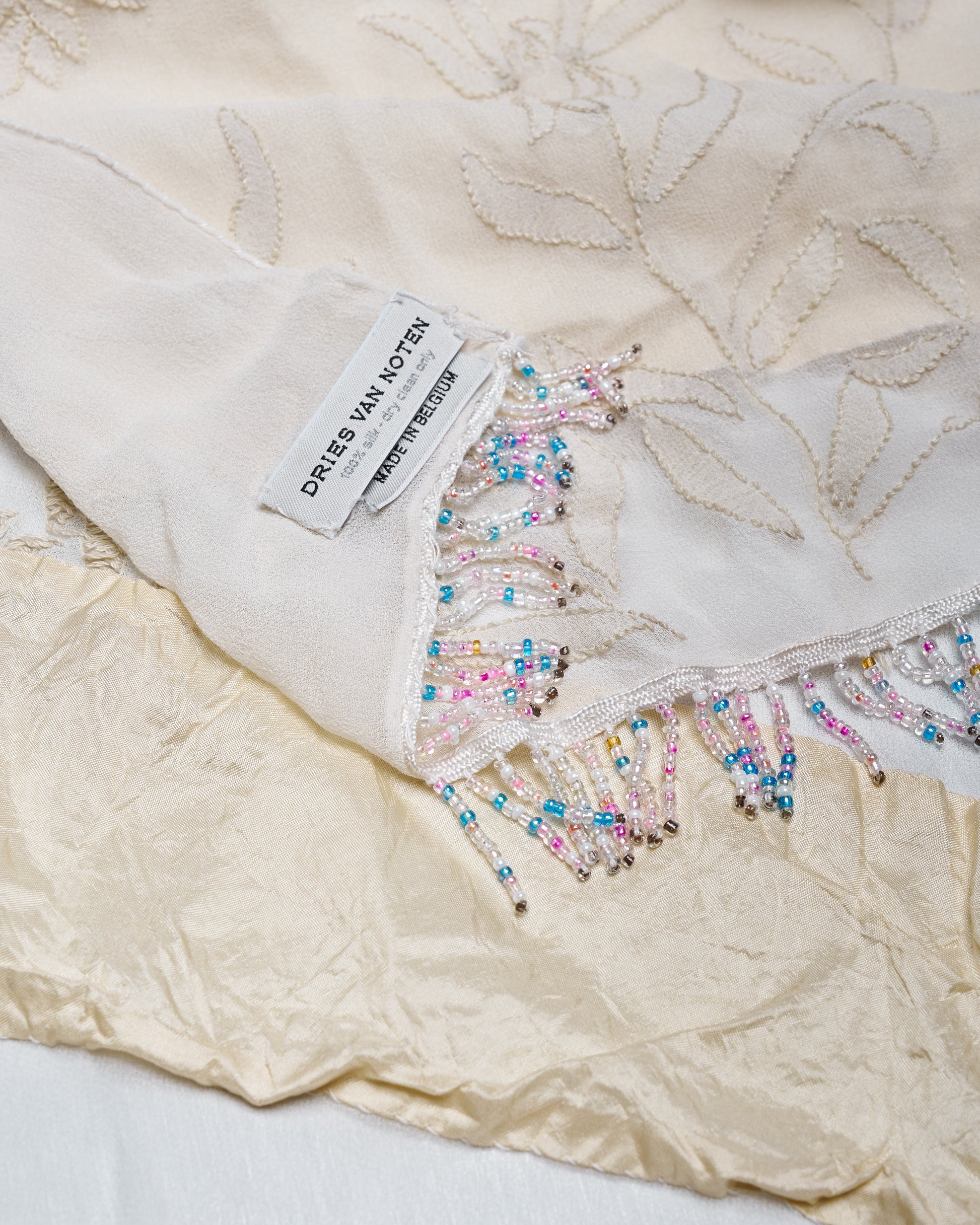 Close-up of a delicate beige fabric with floral embroidery and colorful beaded fringe. A label reads "DRIES VAN NOTEN" indicating luxury fashion.
