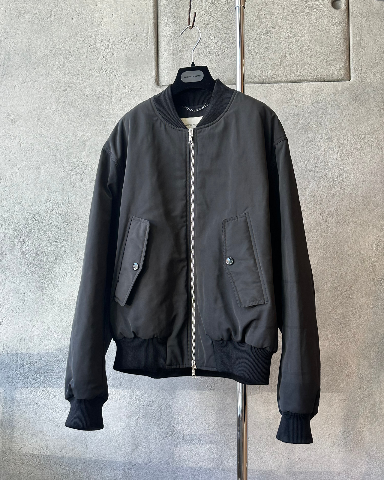 Black Dries Van Noten bomber jacket on a hanger against a light gray wall.