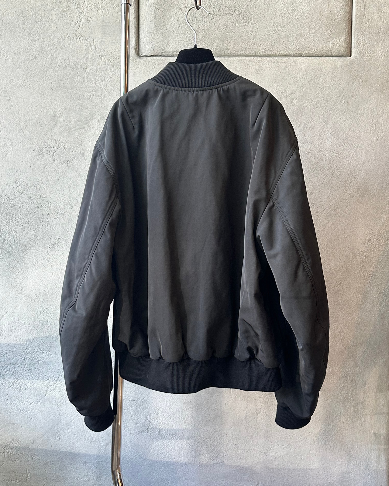 Black Dries Van Noten bomber jacket on a hanger against a light gray wall.