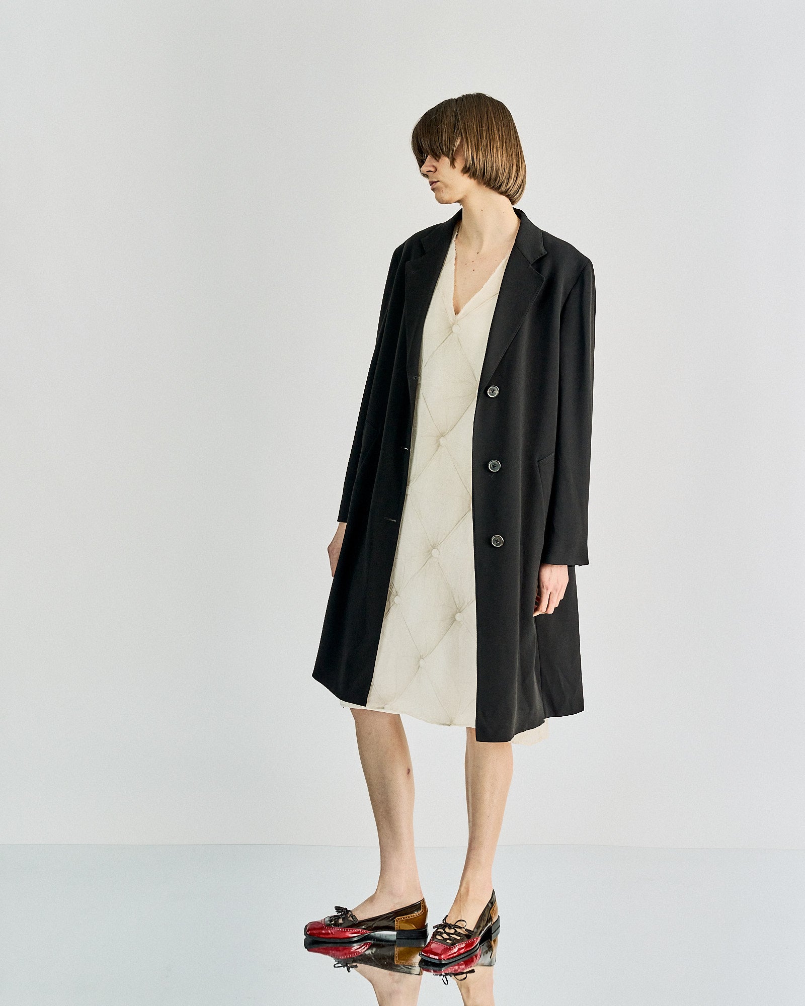 A person with short hair wearing a long black Maison Margiela coat over a white dress, standing against a plain backdrop. They wear red and black shoes, exuding a minimalist and stylish tone.