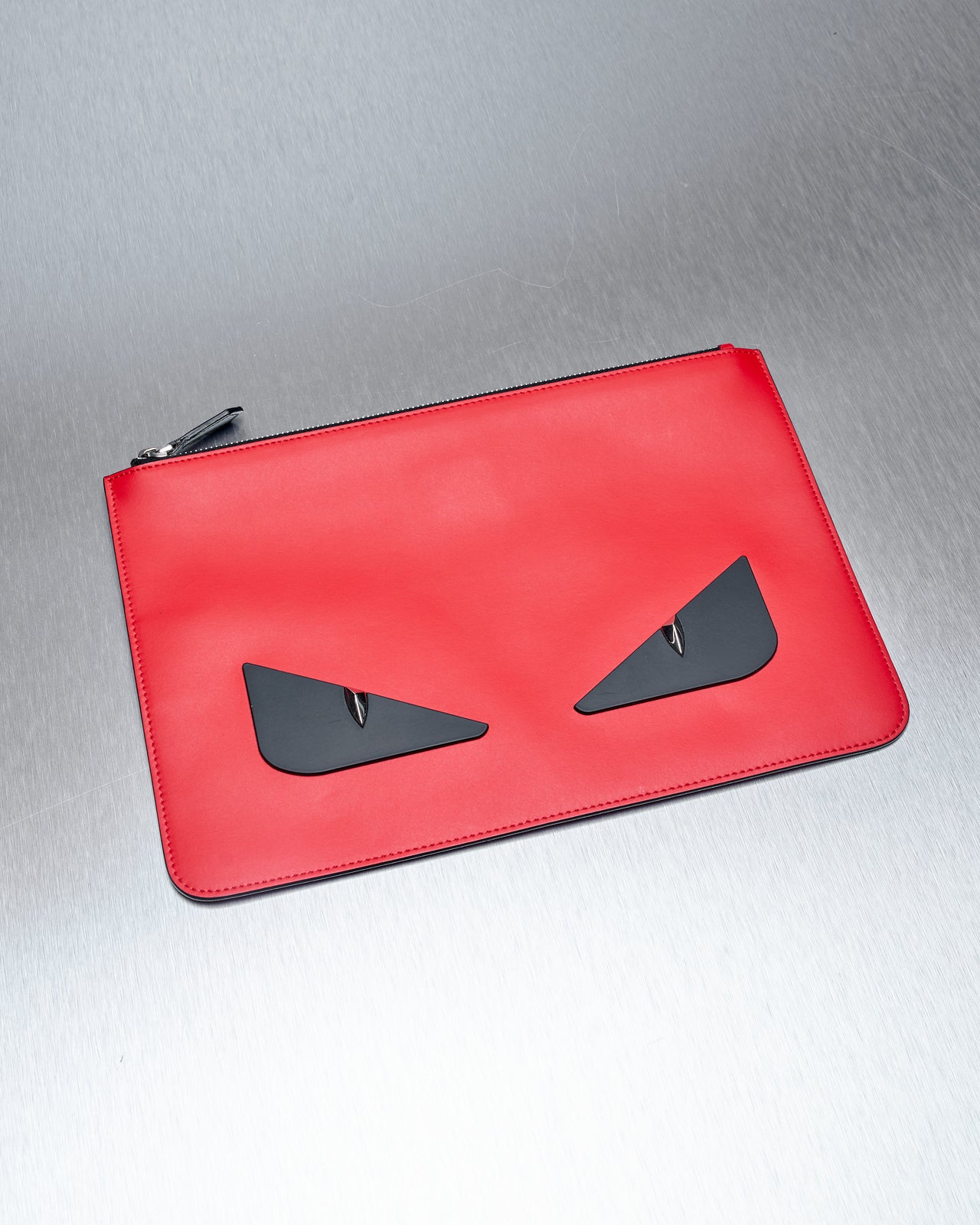 A flat leather Fendi clutch with a silver top-zip and bold black eyes with silver-studded pupils on a reflective surface.