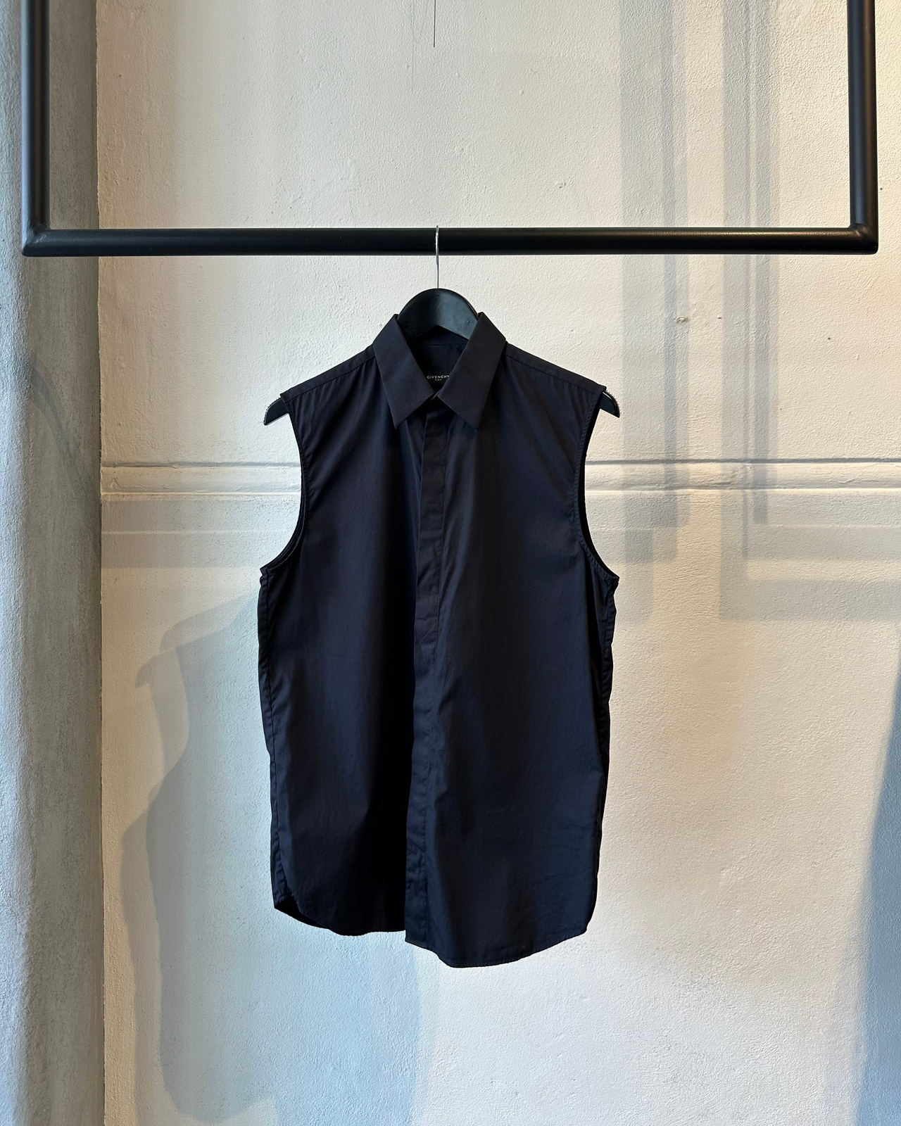 Black sleeveless Givenchy shirt hanging on a rack against a neutral wall.