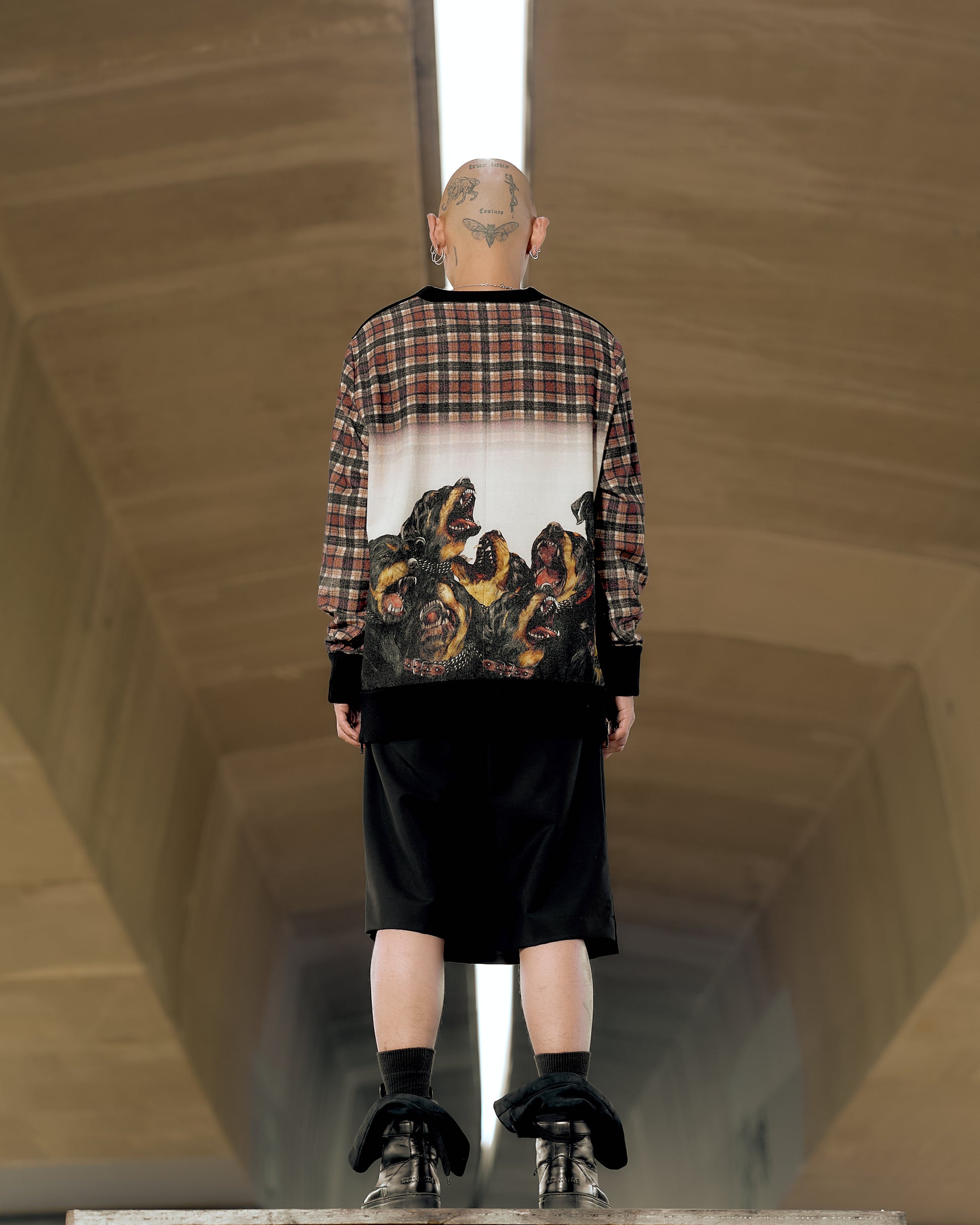 Bald person with tattoos wearing a plaid Givenchy sweater featuring roaring Rottweiler dog faces, black shorts, and boots, standing in a tunnel with an industrial feel.