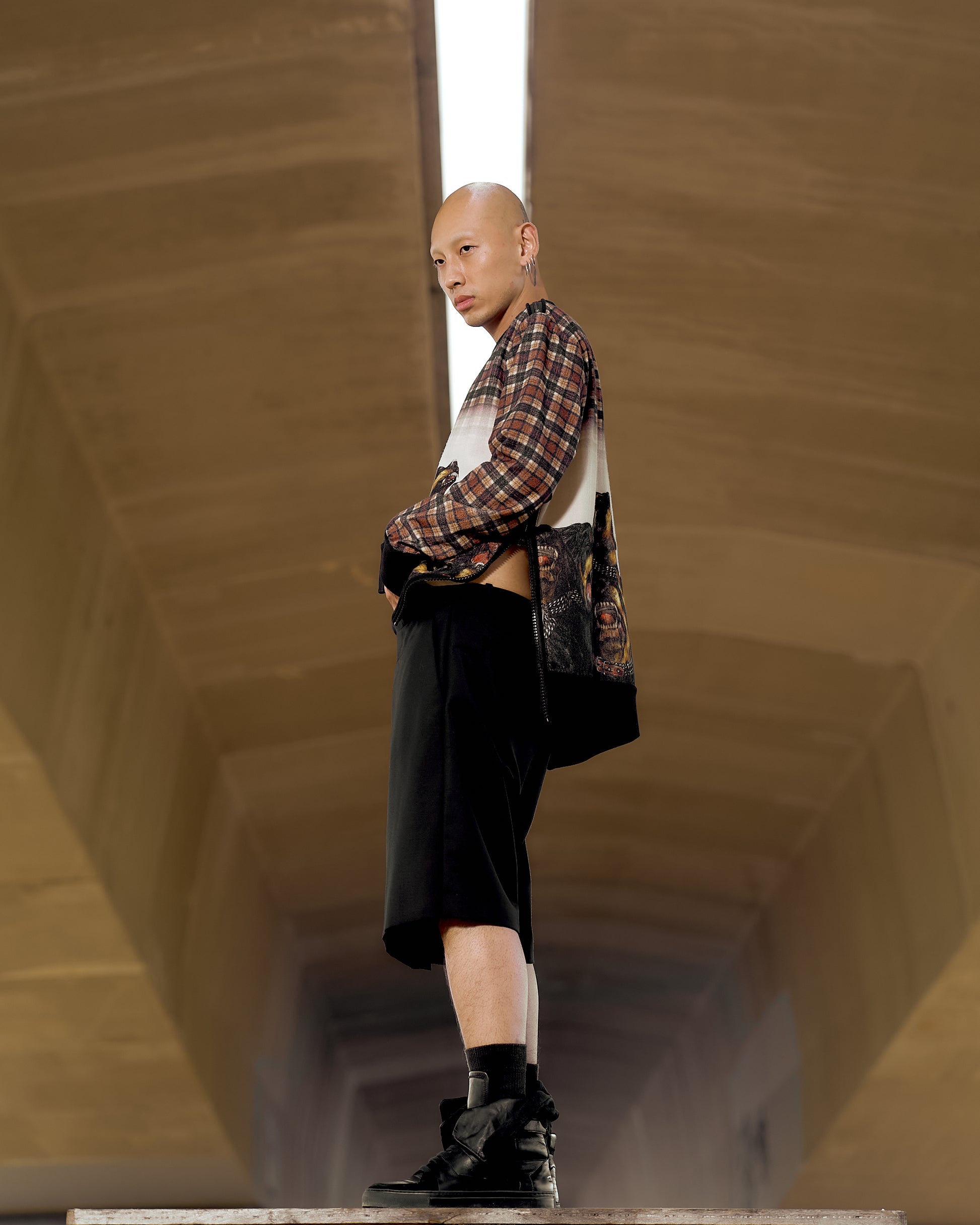 A person stands confidently in a geometric hallway, wearing a checkered and graphic Givenchy sweater with black shorts and boots. The tone is bold and modern.