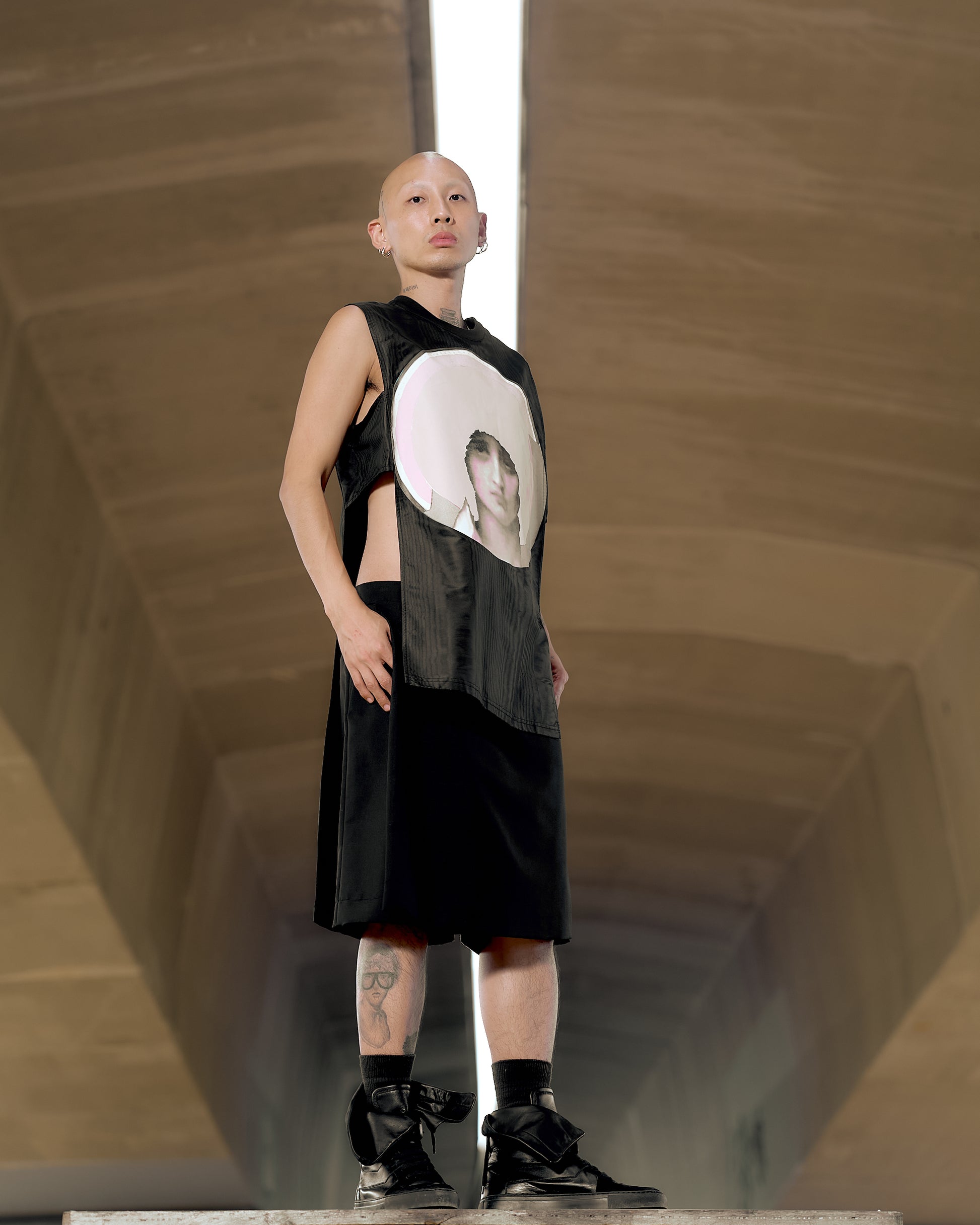 A bald model stands confidently in a modern hallway, wearing a sleeveless black Givenchy outfit with a portrait print.
