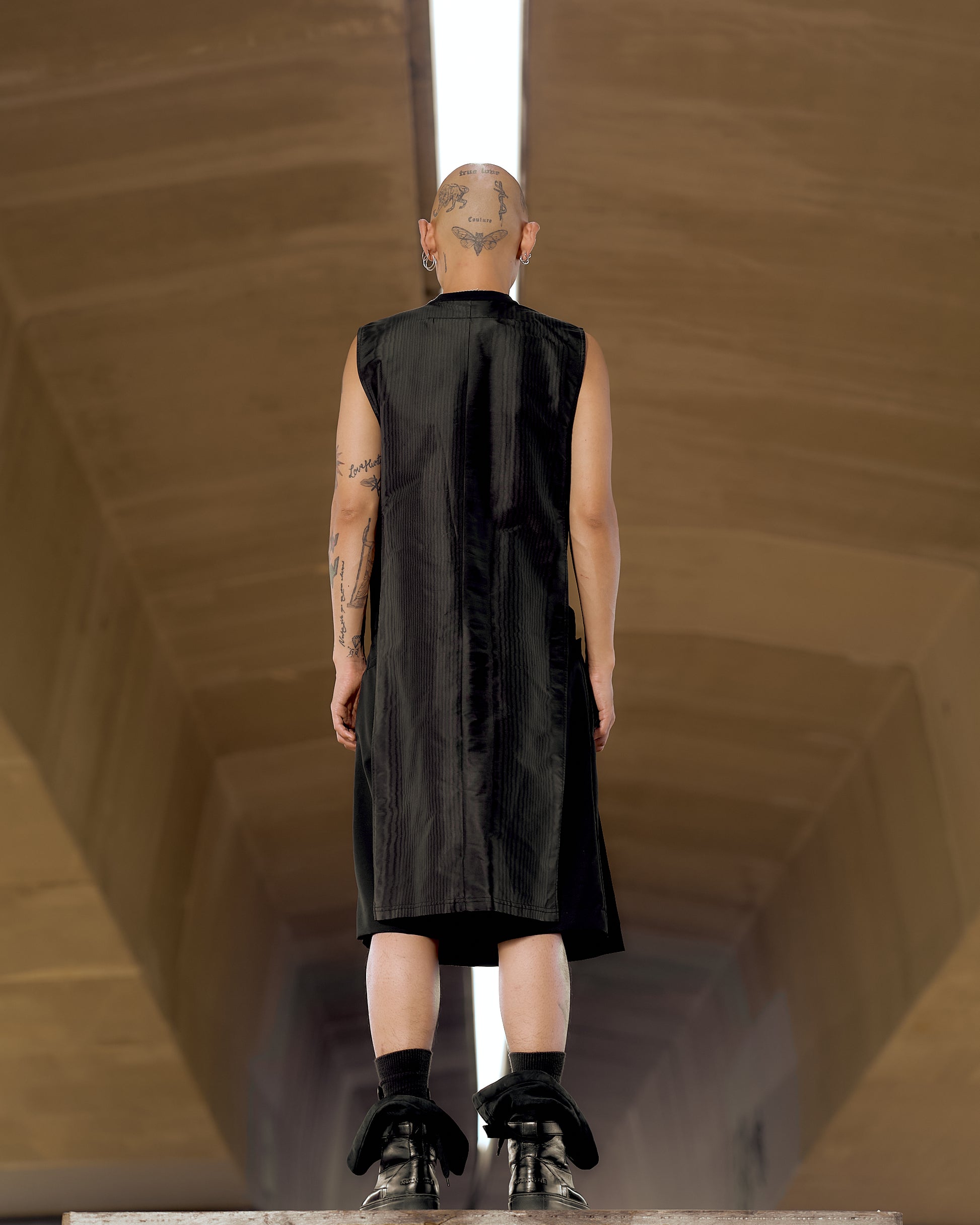 A person stands with their back to the camera, wearing a black sleeveless Givenchy dress and boots. Tattoos are visible on the shaved head and arms. 