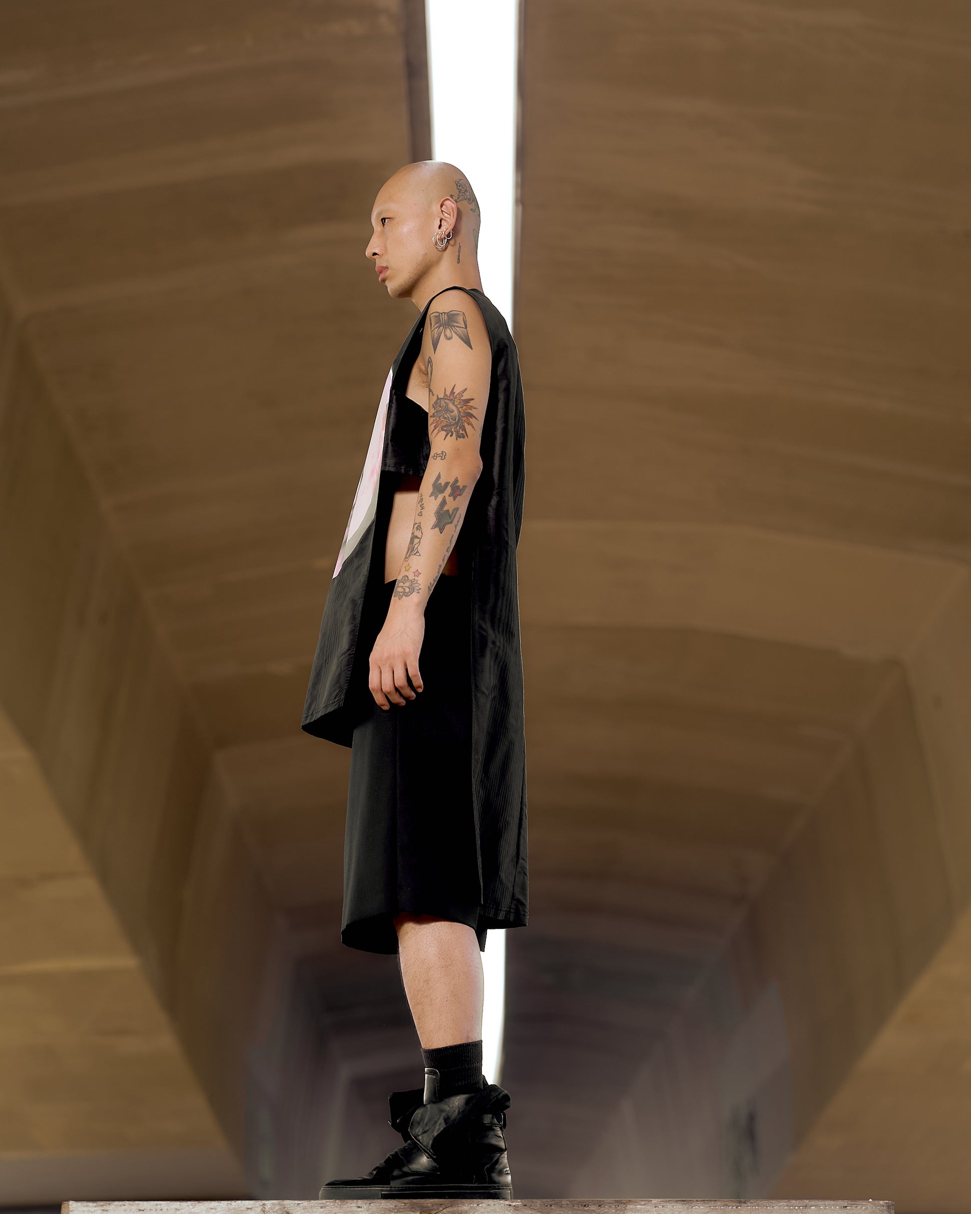 A bald person with tattoos stands in profile under a high ceiling, wearing a sleeveless black Givenchy outfit and boots.