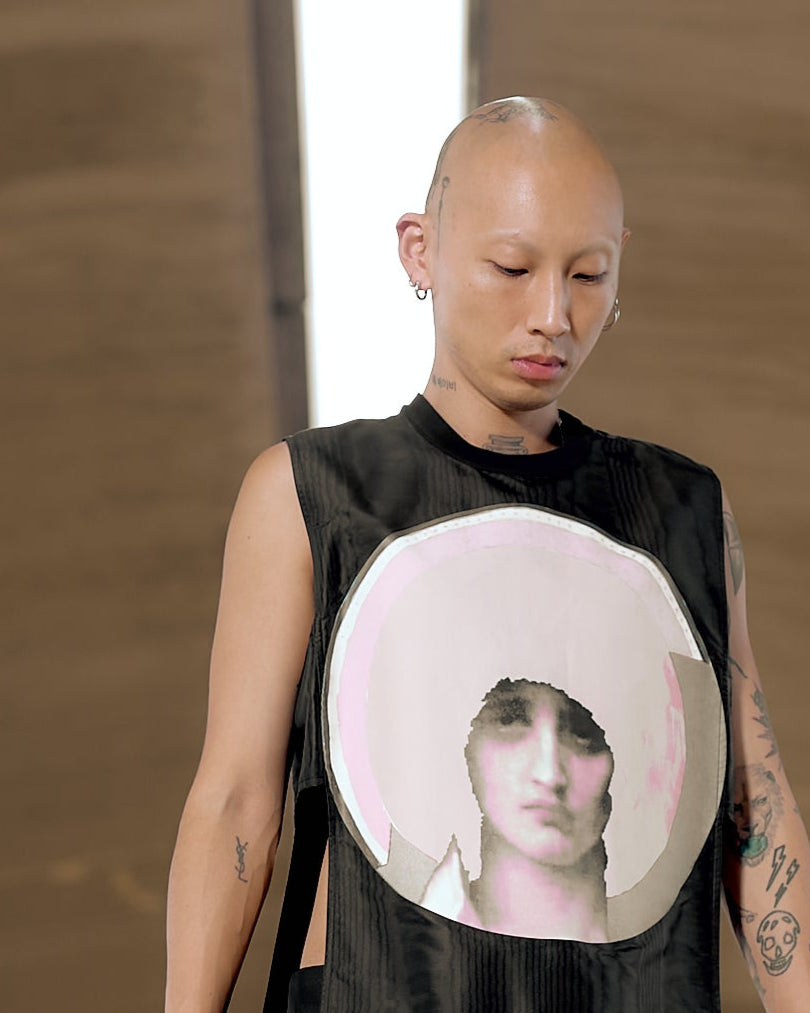 A person with a bald head and earrings is wearing a sleeveless Givenchy top featuring a large abstract face design.