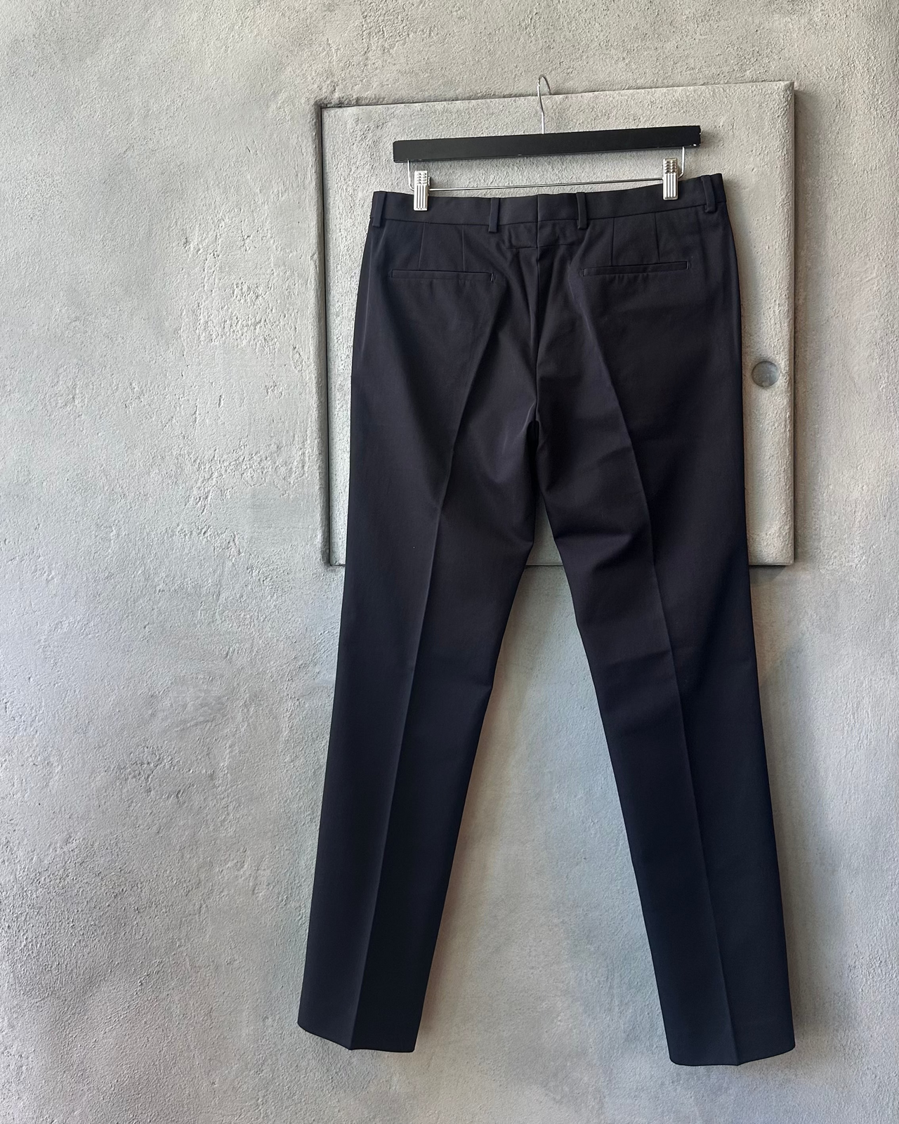 Black Givenchy pants hanging on a rack against a gray concrete wall.