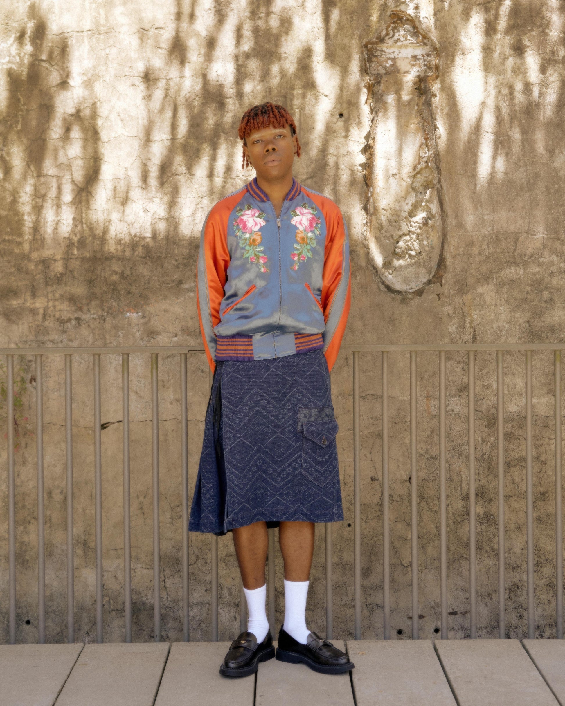 A person stands confidently in front of a textured wall, wearing a floral Gucci bomber jacket, a knee-length patterned skirt, white socks, and black loafers. 