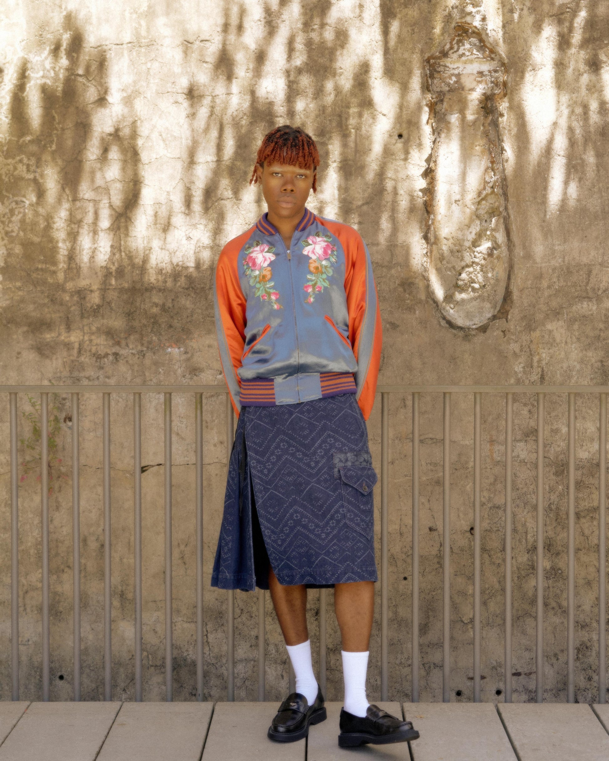A person stands confidently in front of a textured wall, wearing a floral Gucci bomber jacket, a knee-length patterned skirt, white socks, and black loafers. 