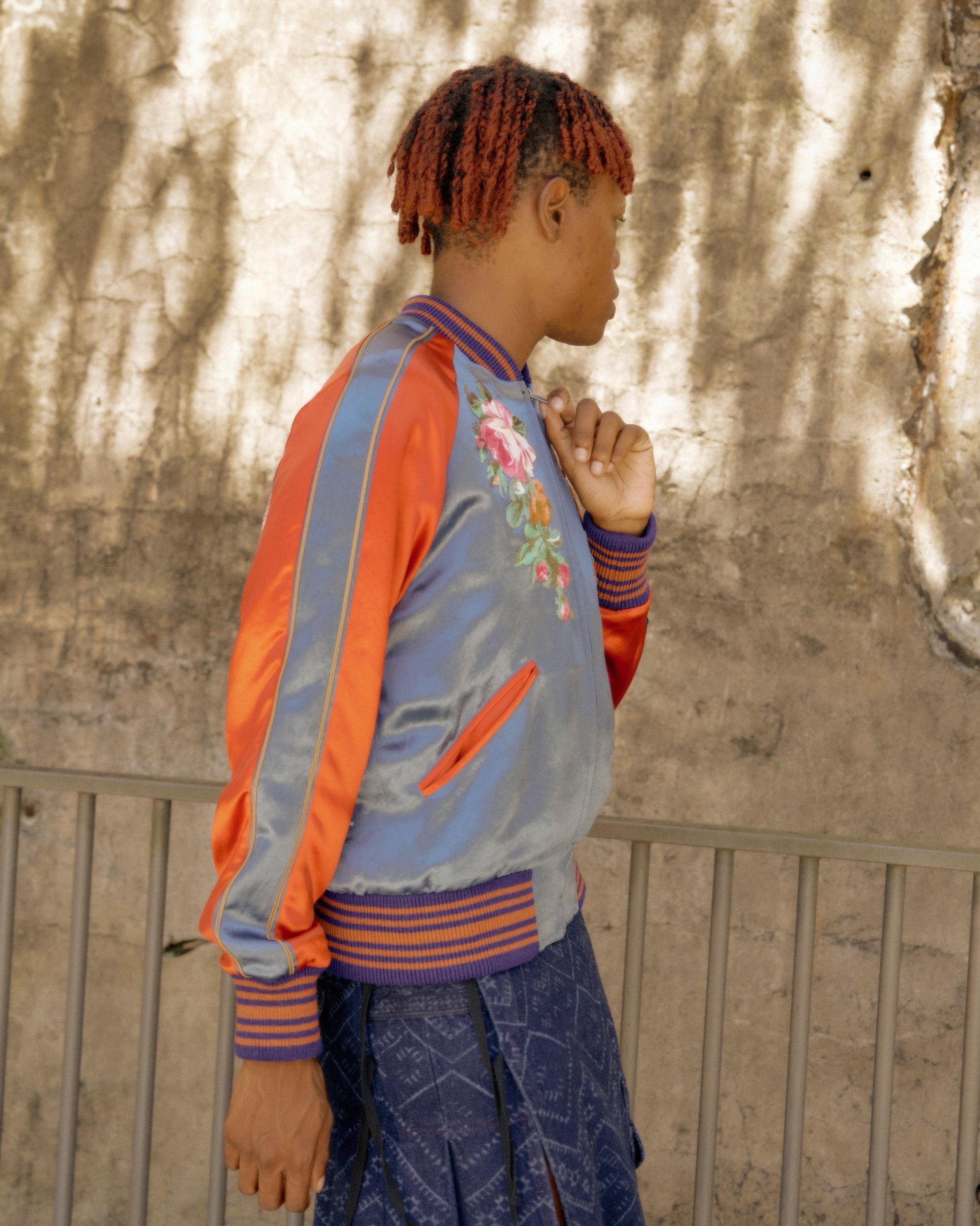 Person with short, red hair stands sideways wearing a blue and orange Gucci satin jacket with floral embroidery. Background shows a textured, sunlit wall.