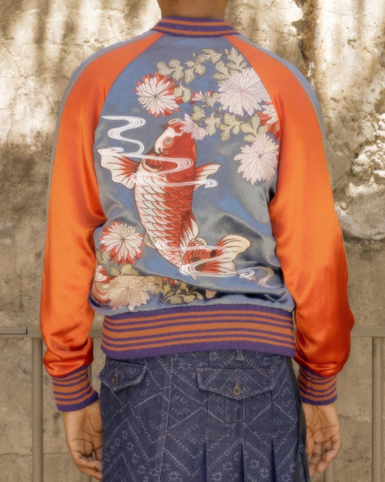 Back view of a person wearing a vibrant Gucci jacket with a koi fish and floral design. The jacket features bold orange and blue hues, conveying a lively tone.