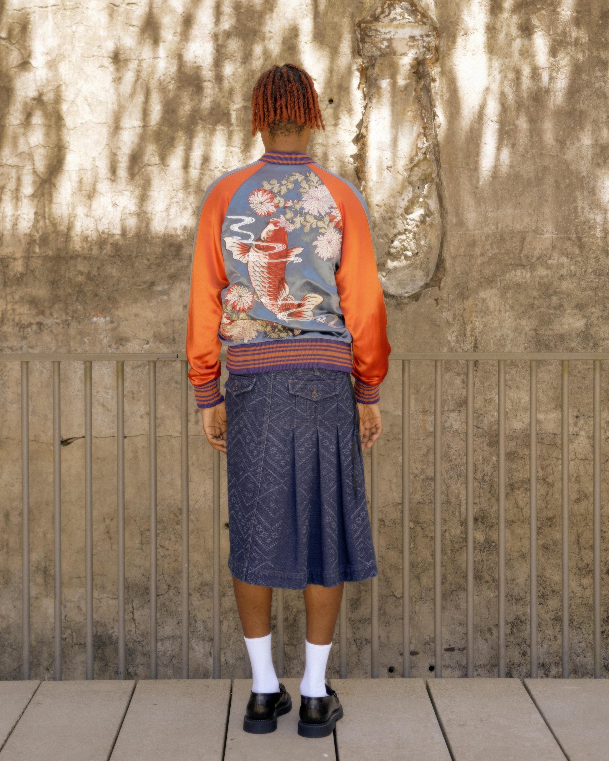 A person stands with their back to the camera, wearing a colourful Gucci bomber jacket featuring a koi fish design and a pleated denim skirt. The wall behind is textured, with shadows cast on it.