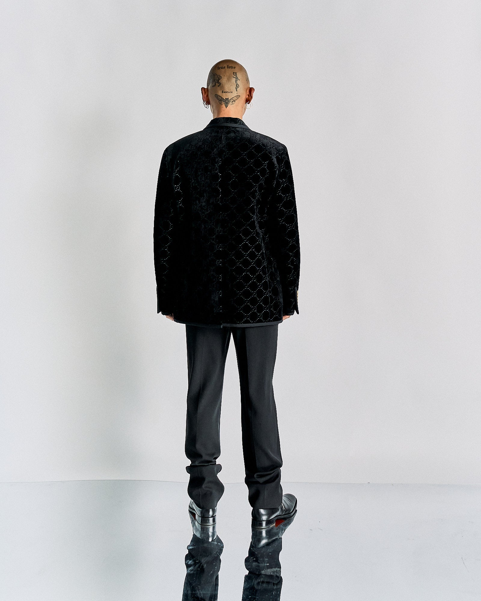 A person stands facing away, wearing a textured black Gucci suit and shiny shoes on a reflective floor. 