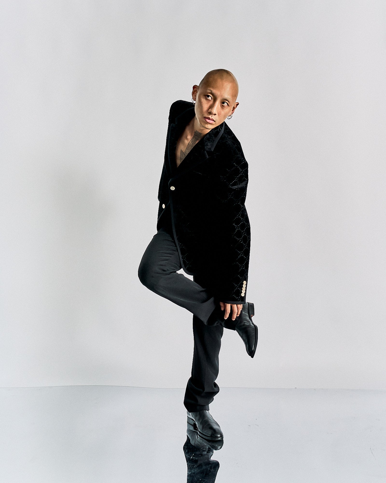 A person with a shaved head poses against a plain backdrop, wearing a textured black Gucci coat and black pants.