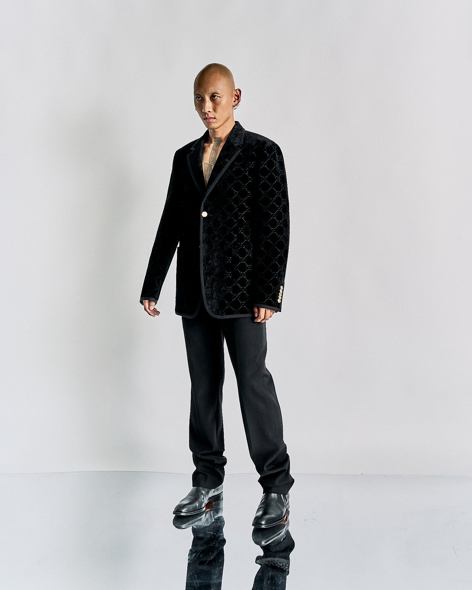 A person stands confidently against a plain background, wearing a textured black Gucci blazer, black pants, and shiny black shoes. 