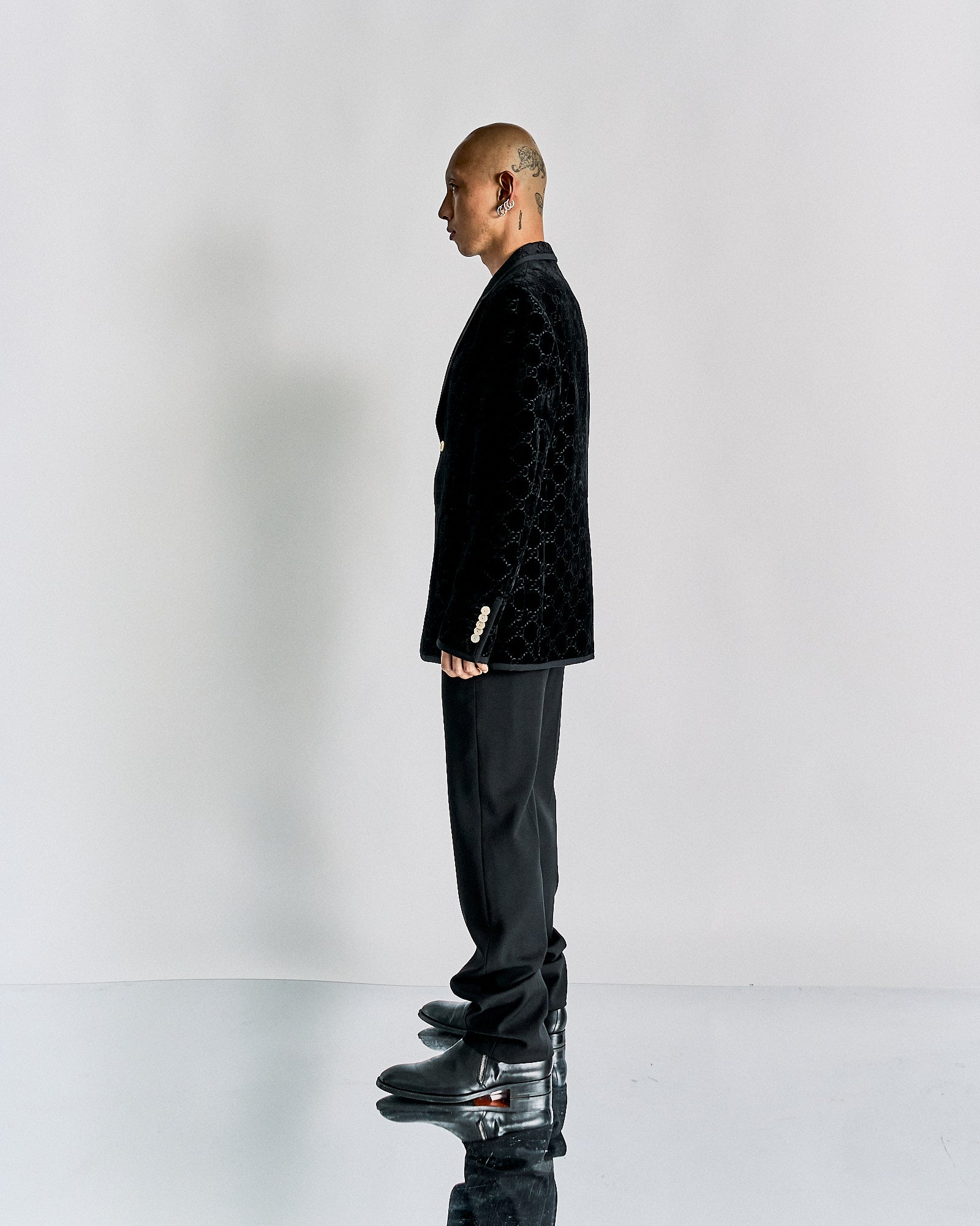 A person stands in profile against a plain backdrop, wearing a black Gucci velvet jacket, black pants, and shiny black shoes.