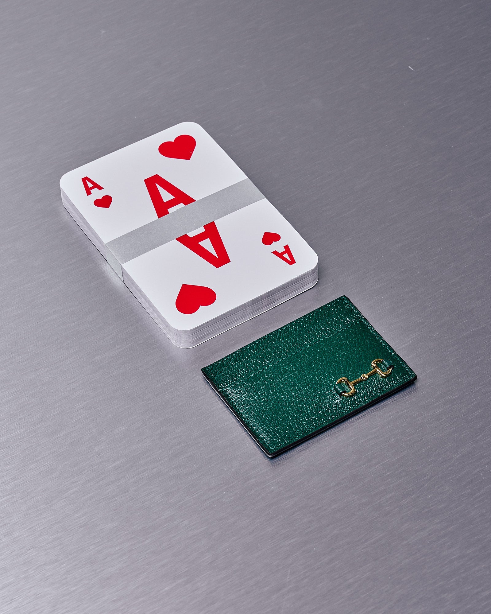 A large deck of Ace of Hearts Gucci playing cards sits beside a small green leather cardholder with gold detailing, on a metallic surface.
