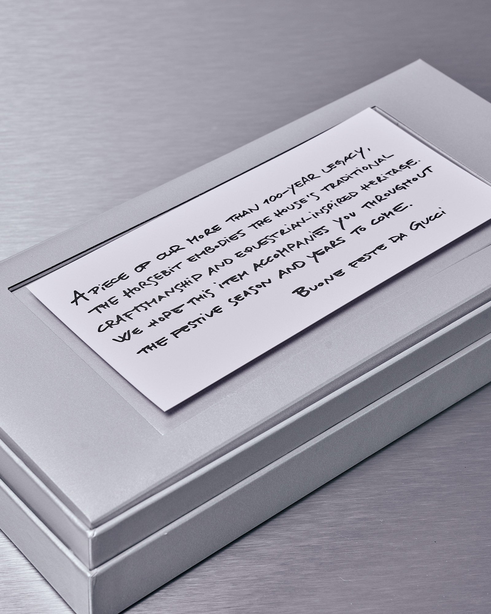 A silver Gucci box on a metallic surface features a handwritten card. The message speaks of craftsmanship and heritage