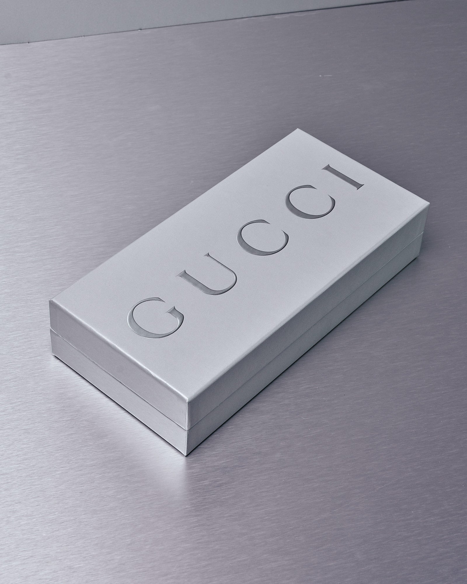 A sleek, gray gift box on a metallic surface features the word "GUCCI" embossed in large, bold letters, conveying luxury and elegance.