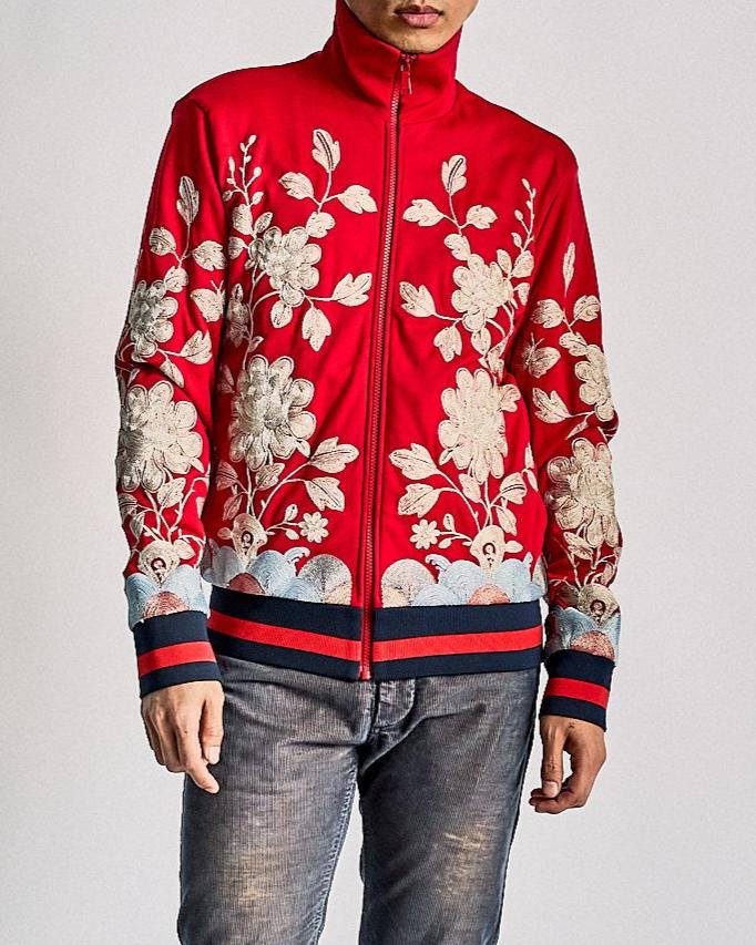 A person wearing a red Gucci jacket with prominent floral embroidery in white and light blue. The jacket has striped cuffs and waistband, paired with faded jeans.