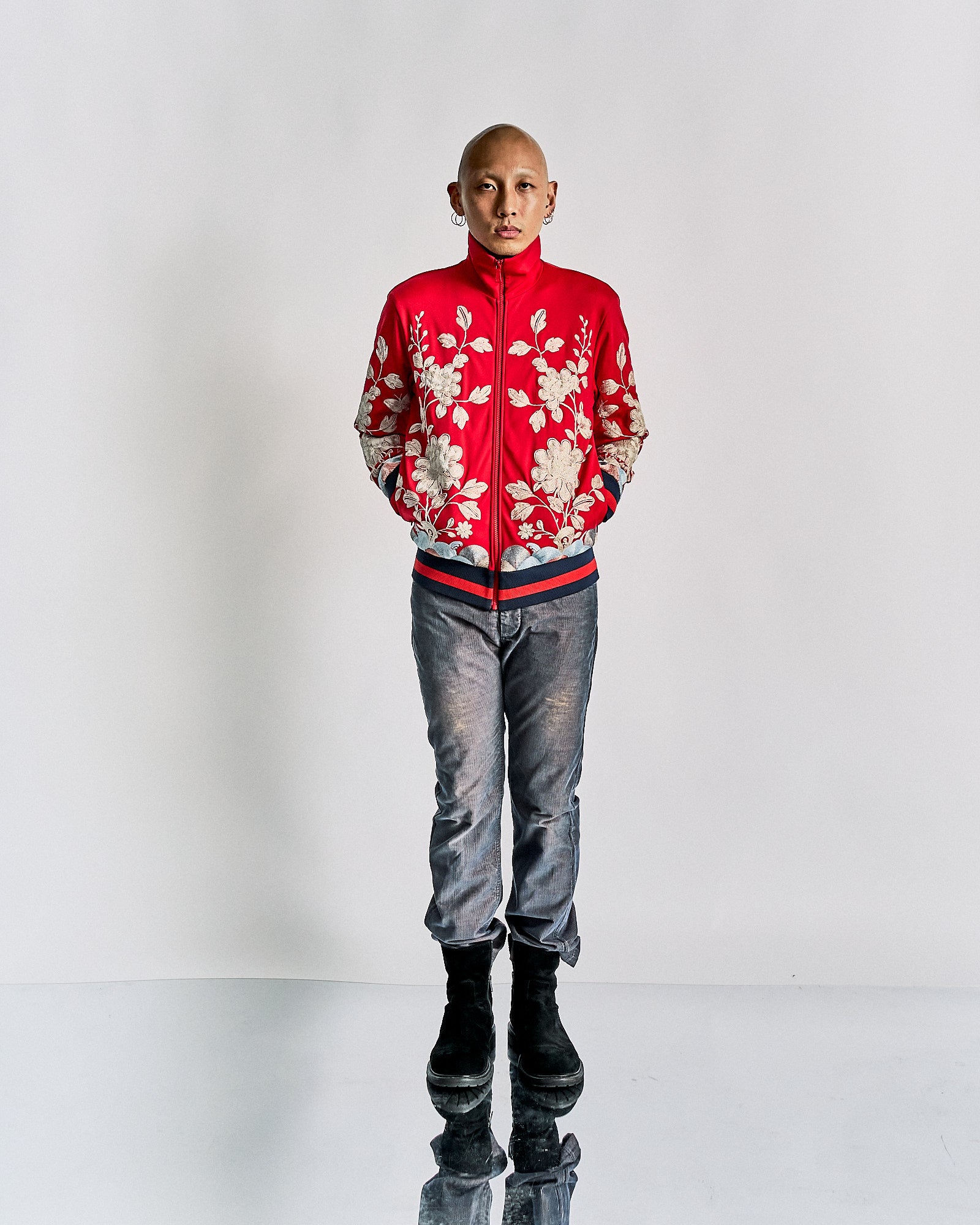 A person stands confidently against a white background, wearing a vibrant red Gucci jacket with floral designs, blue jeans, and black boots. 