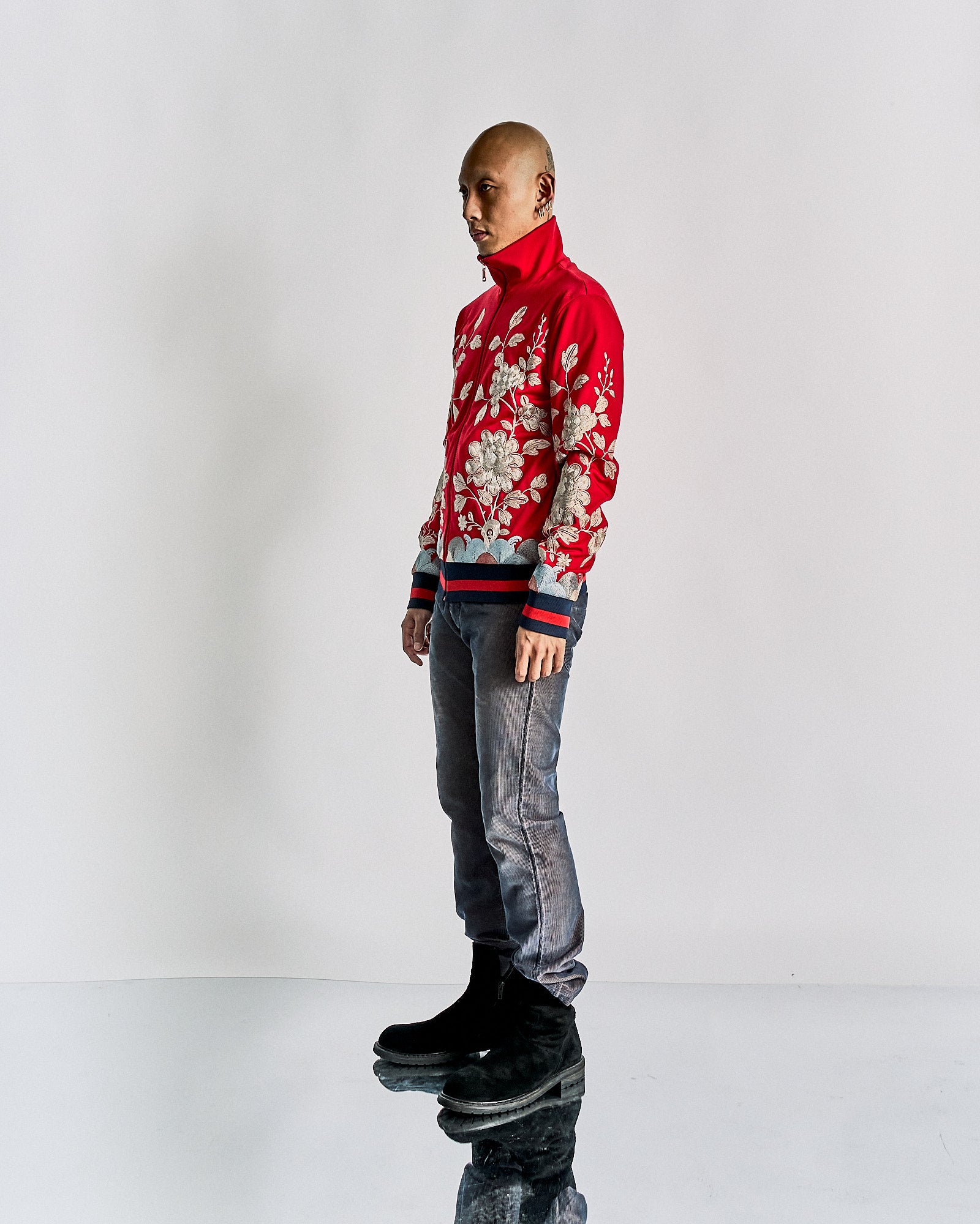 A person stands against a white background, wearing a bold red Gucci jacket with intricate floral patterns, distressed jeans, and black boots, exuding a stylish and modern vibe.