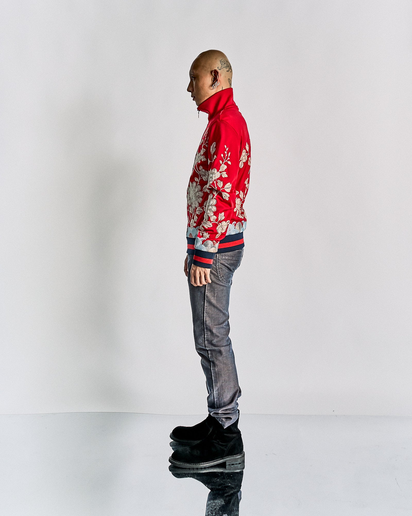 Side profile of a person standing against a plain background, wearing a red Gucci jacket with white floral patterns, gray jeans, and black boots. 