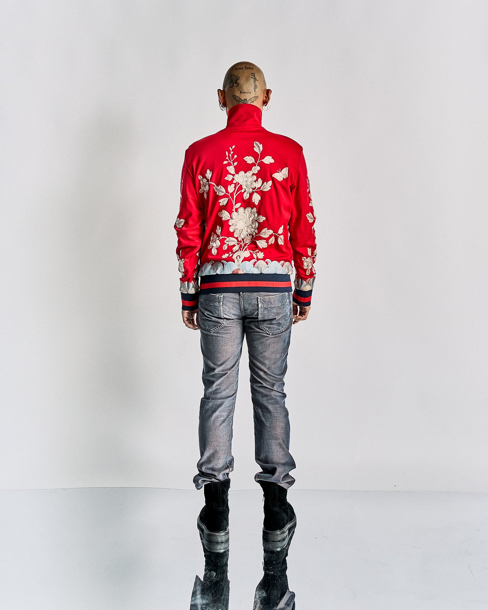 A person stands facing away, wearing a vibrant red Gucci jacket adorned with white floral embroidery. They have short hair and are in jeans and boots against a plain background.
