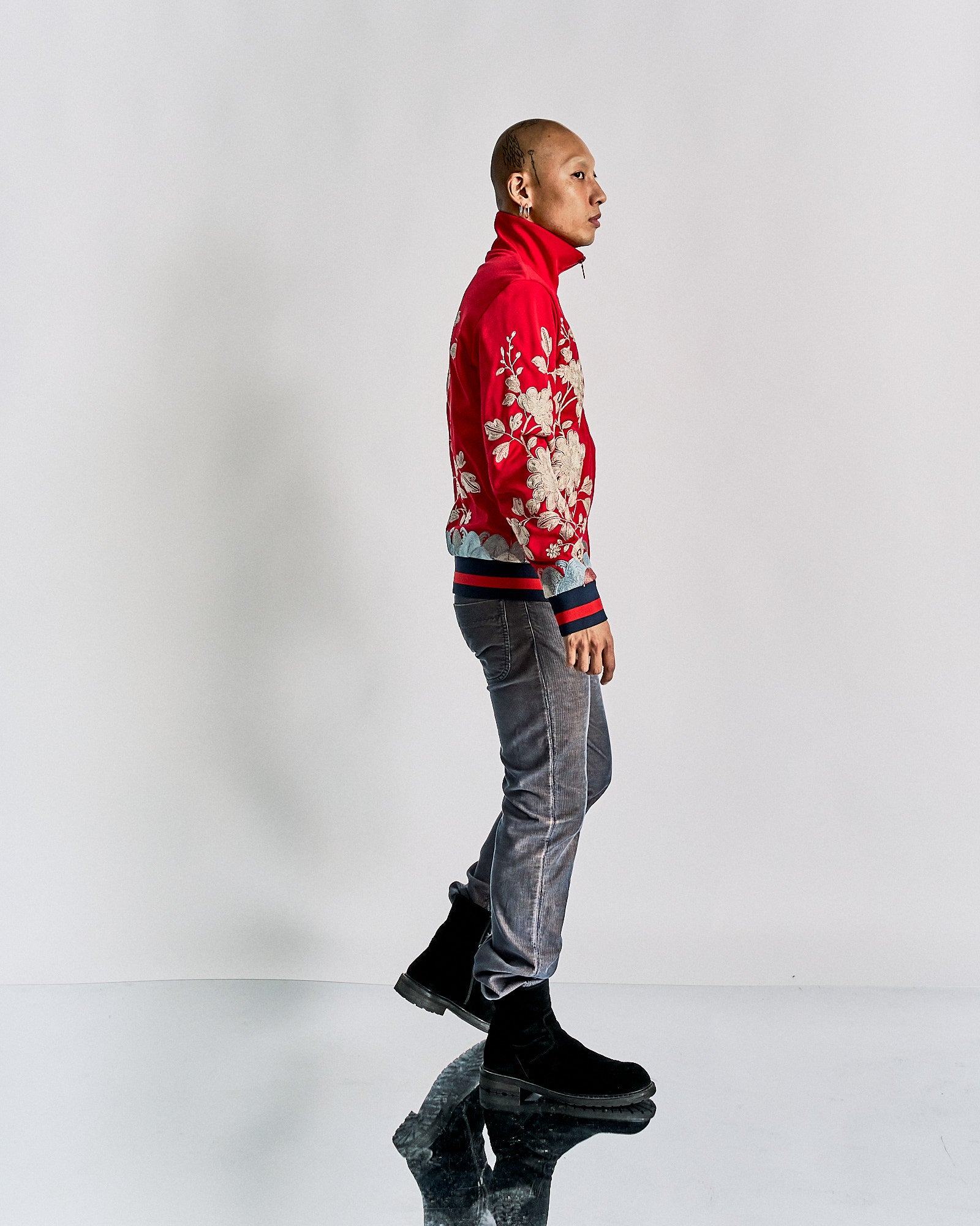 A person stands in profile against a plain backdrop, wearing a red floral Gucci jacket, gray jeans, and black boots