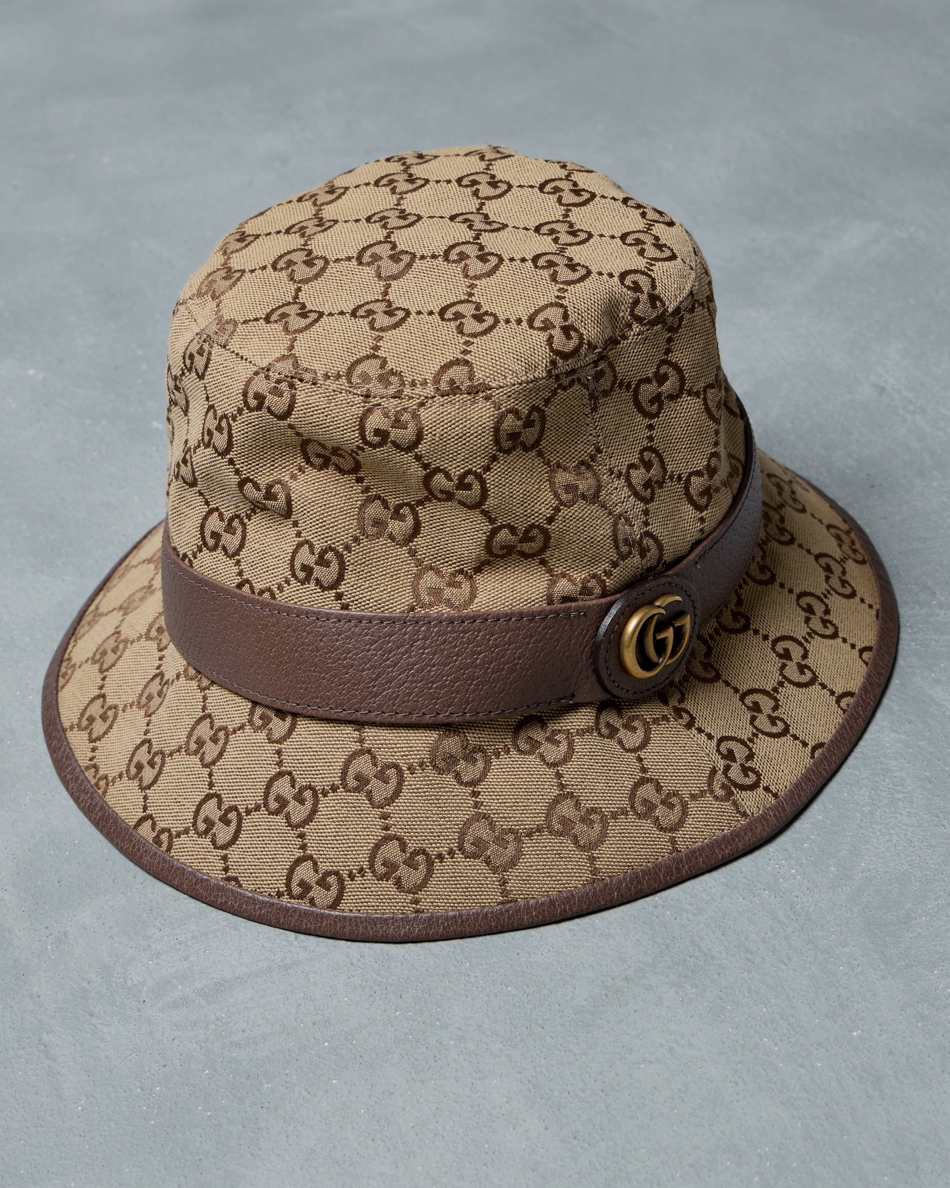 Beige Gucci bucket hat with brown logo pattern and leather band on a gray background