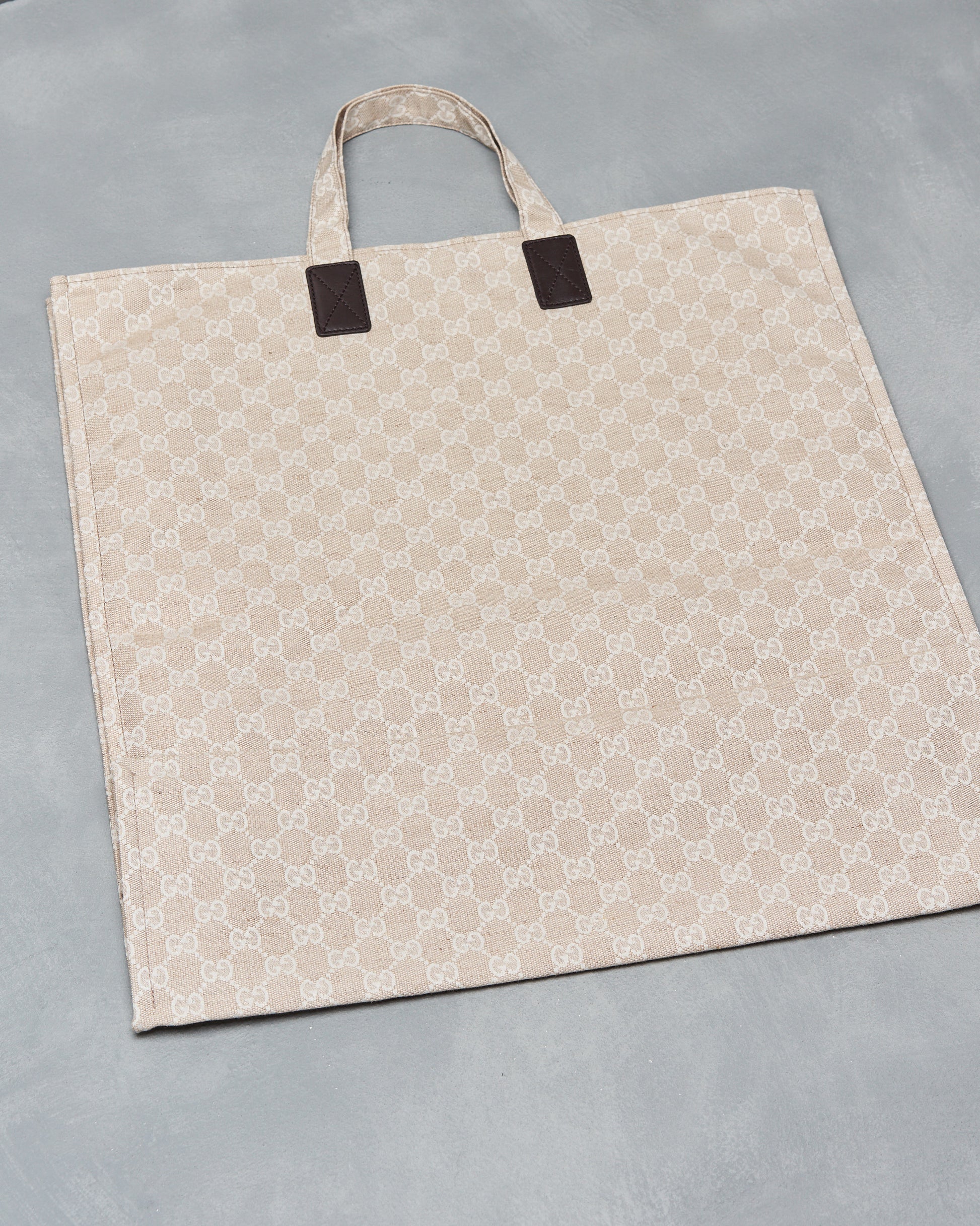 Beige Gucci tote bag with a subtle, repeating monogram pattern, featuring brown leather accents and handles. It rests on a neutral gray surface.