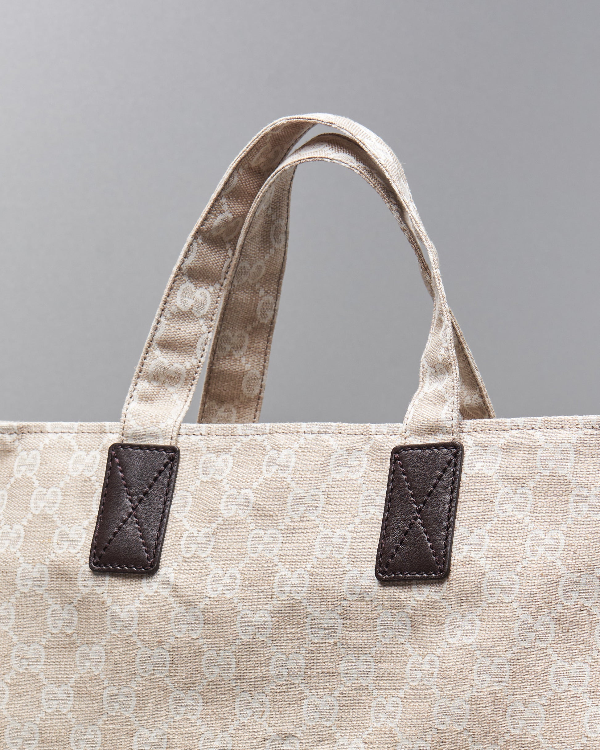 Close-up of a beige Gucci canvas tote bag with a subtle repeating pattern. The handles are attached with dark leather patches, stitching clearly visible.