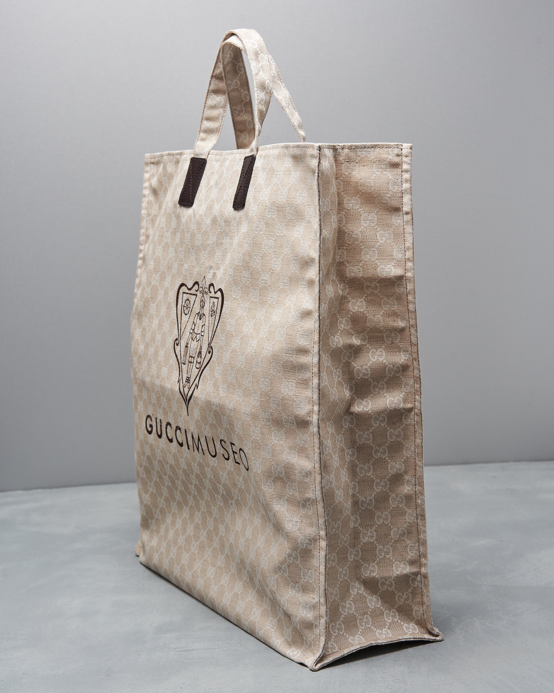 Beige Gucci Museo tote bag with a subtle monogram pattern, brown handles, and an emblem. It rests on a gray surface with a minimalist feel.