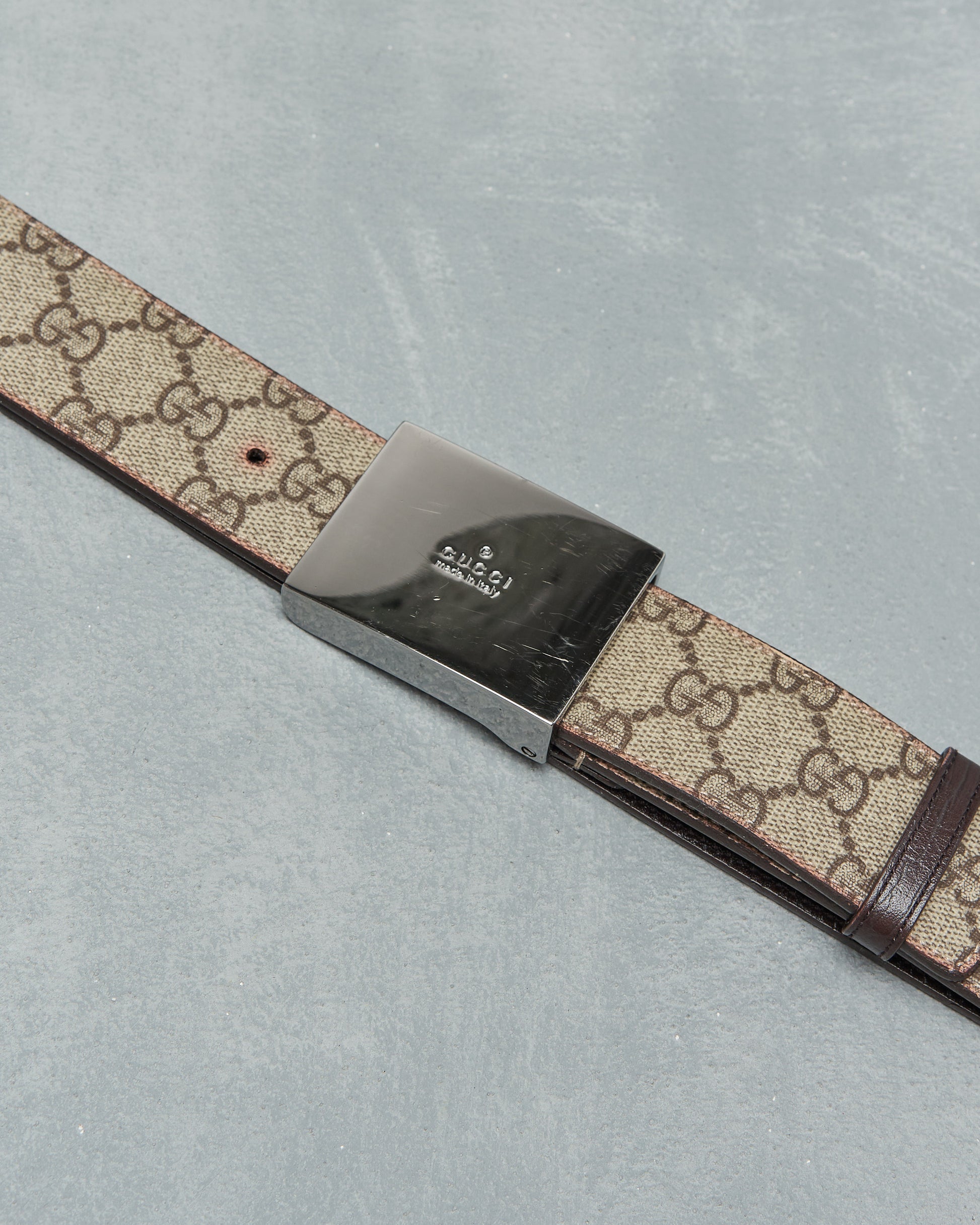 A designer Gucci belt with a silver buckle and beige strap featuring a repeating pattern of interlocking logos. The background is a textured grey surface.