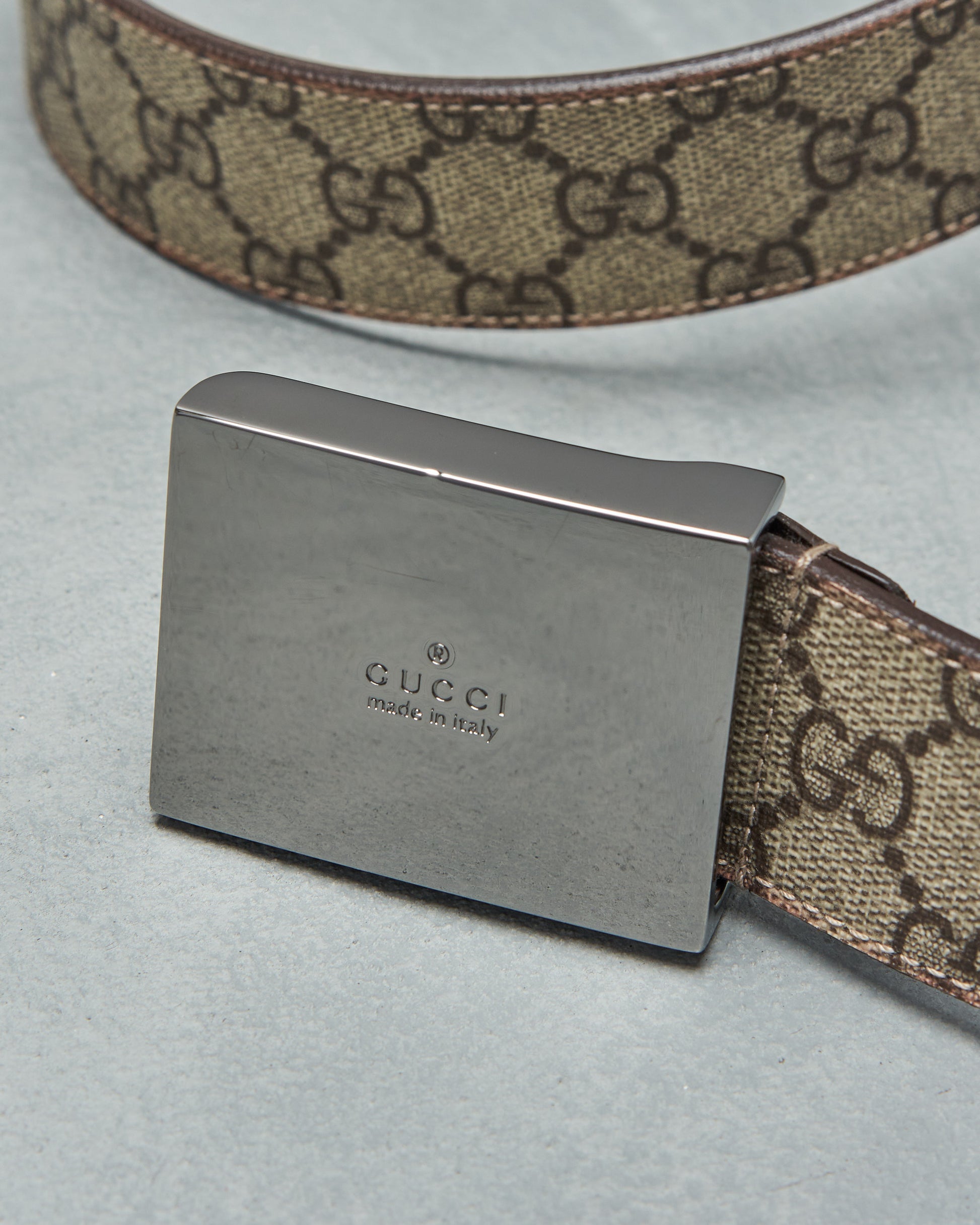 Close-up of a designer belt featuring a metal buckle with "Gucci, made in Italy" engraving. The fabric strap is brown with a repeating GG pattern.