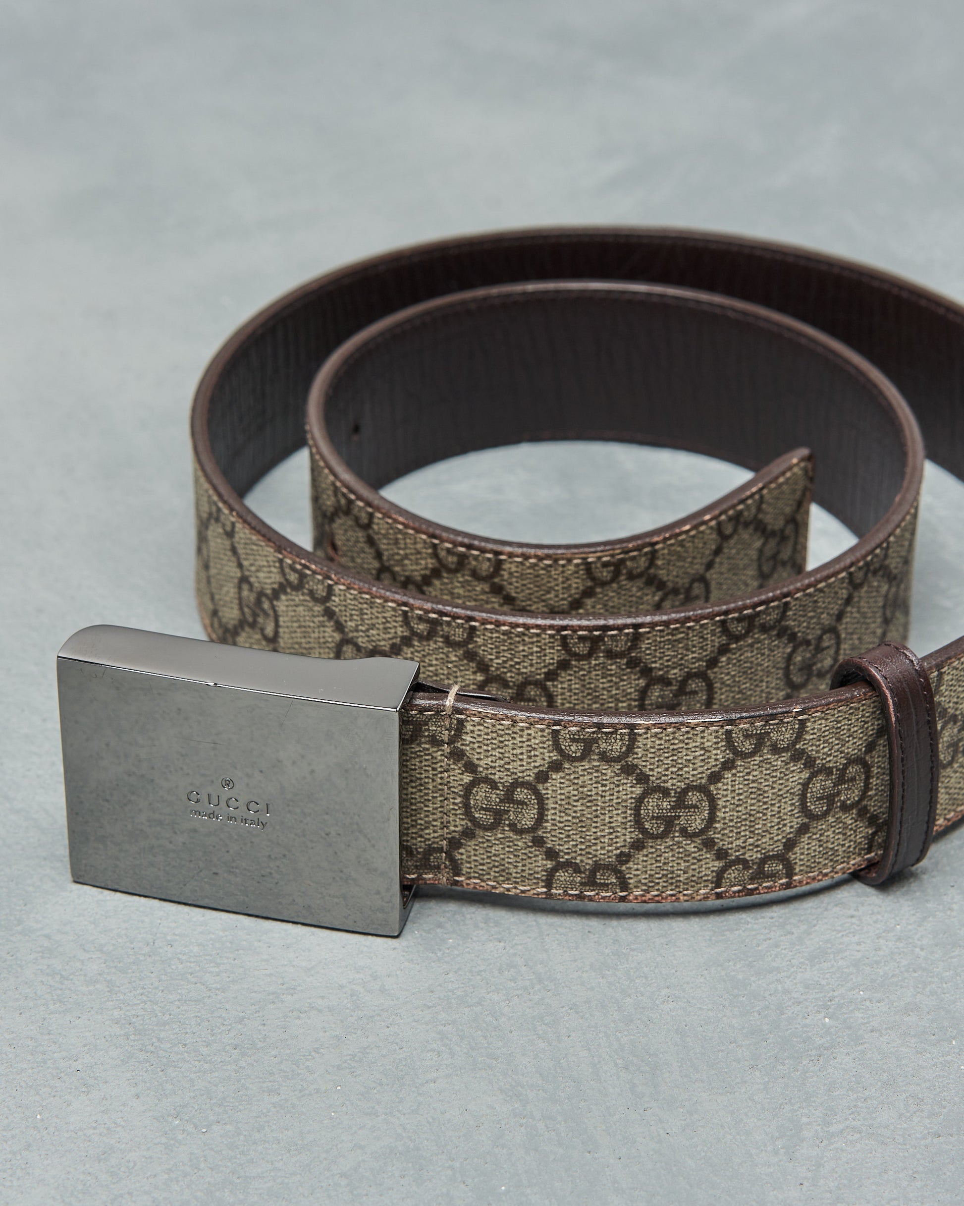 A brown leather Gucci belt with a polished metal buckle featuring the Gucci GG Supreme logo embossed on it.
