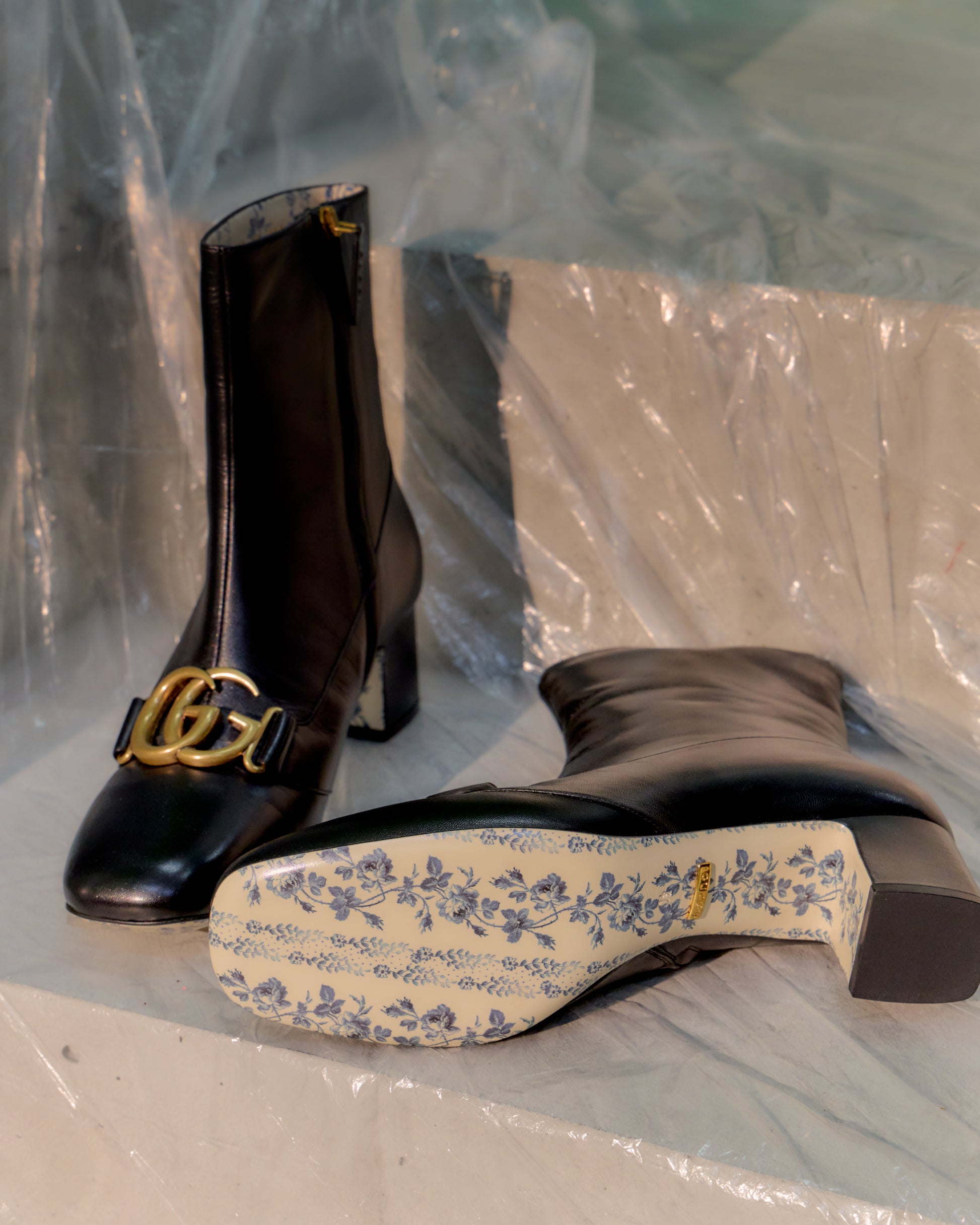 Black high-heeled Gucci boots with gold chain detail on a clear plastic sheet.
