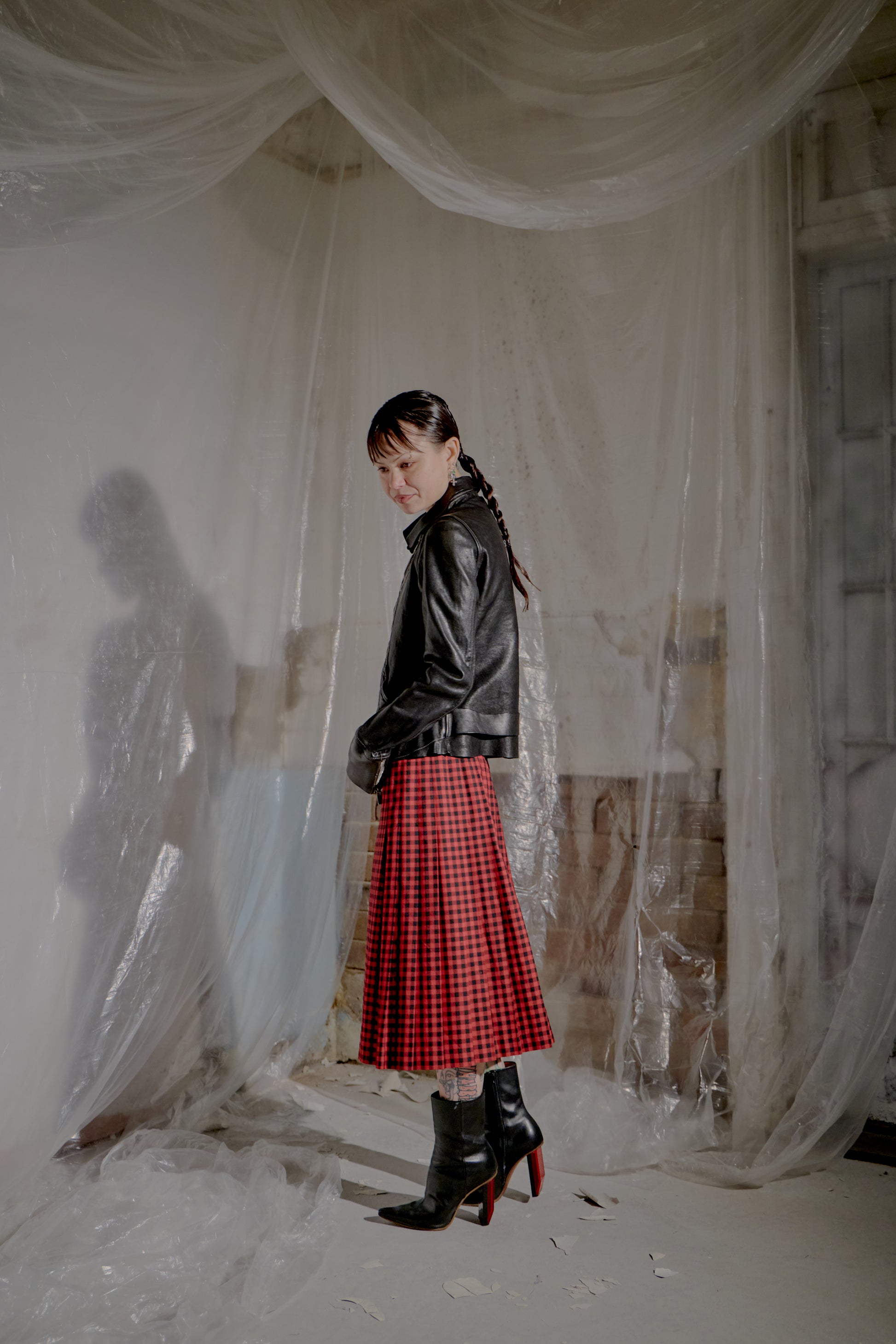 Person wearing a black leather Guidi jacket and red plaid skirt standing in a room with white curtains.