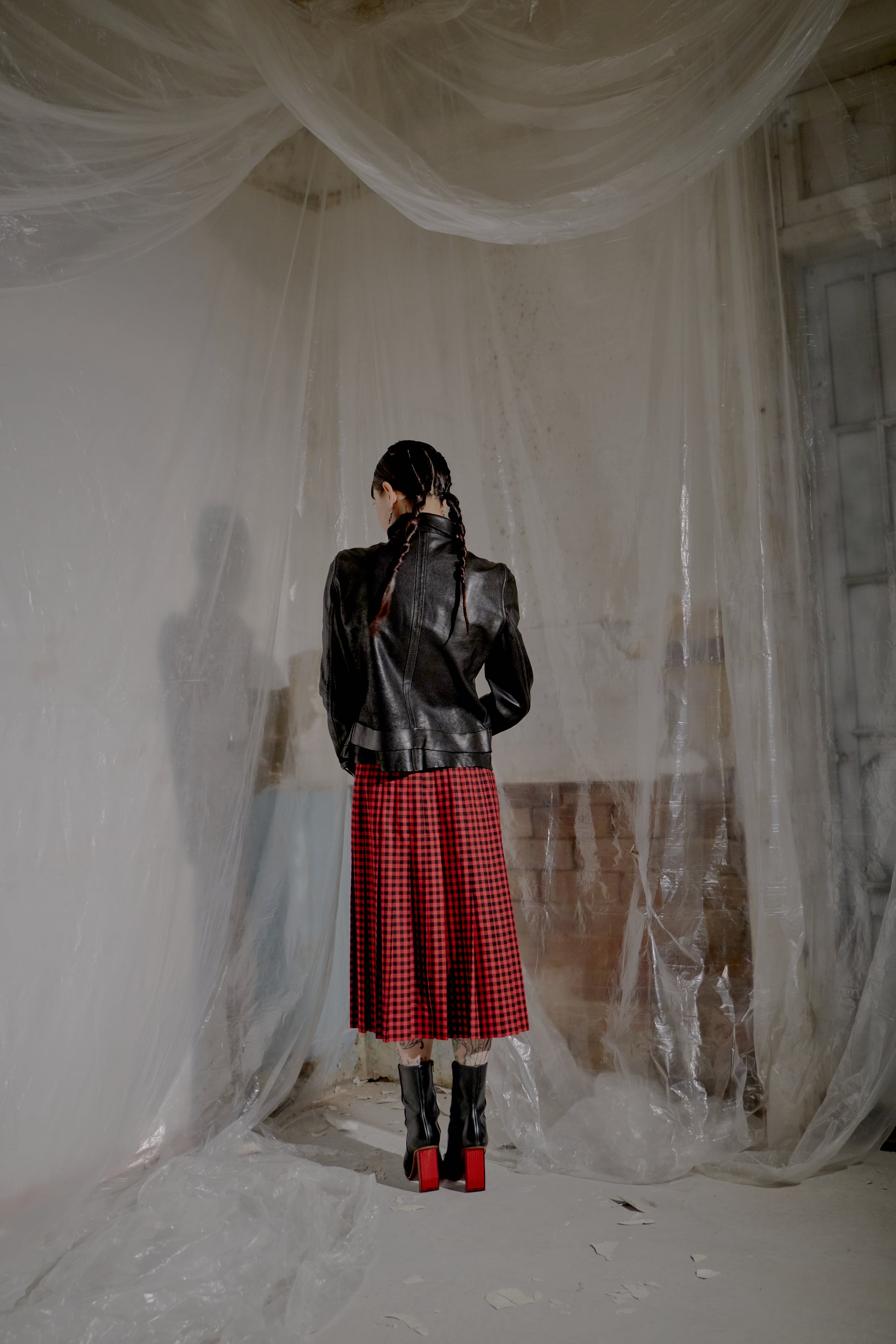 Person wearing a black leather Guidi jacket and red plaid skirt standing in a room with plastic sheeting.
