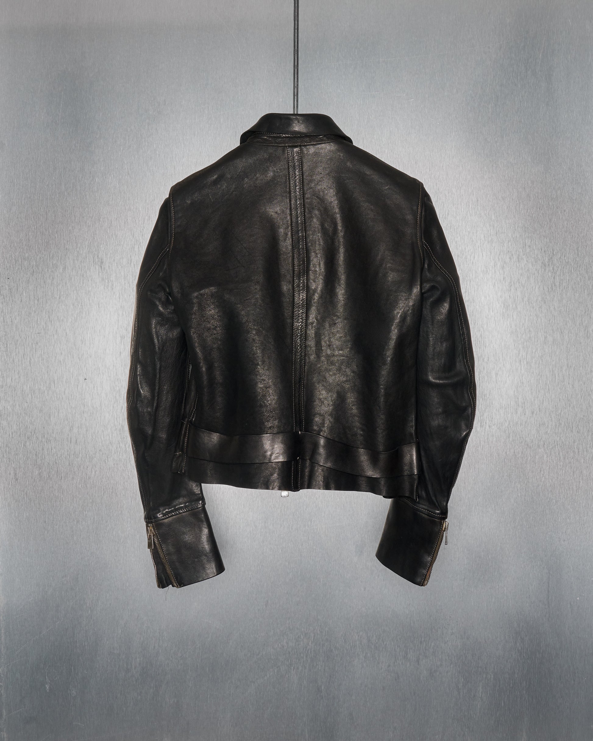 A black leather Guidi jacket hangs against a plain grey background. The jacket's back is visible, showing sleek design and glossy texture, exuding a classic, stylish vibe.