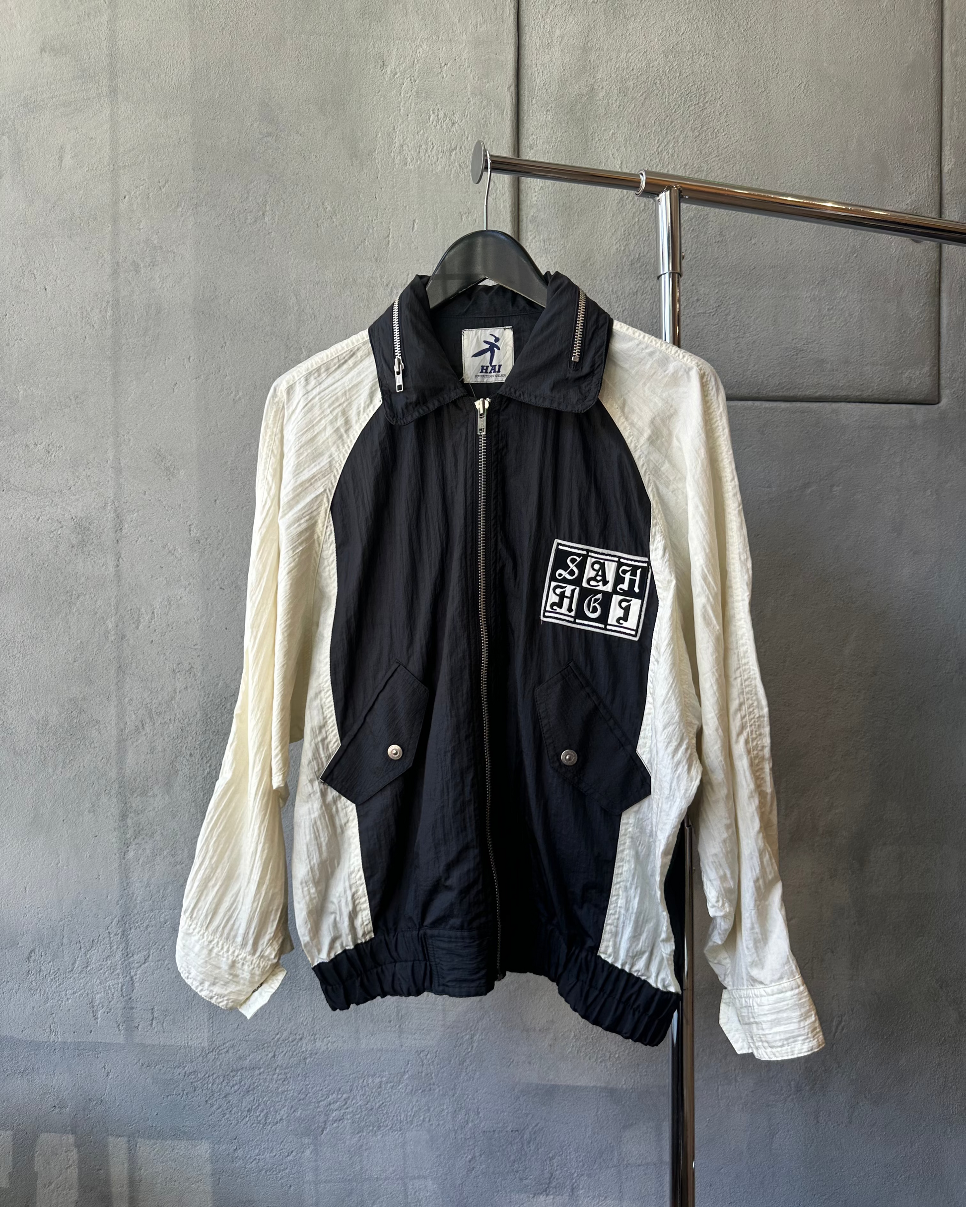 A black and white Hai Sporting Gear jacket on a hanger against a gray wall. It features two front pockets and embroidered text on the chest, exuding a retro vibe.