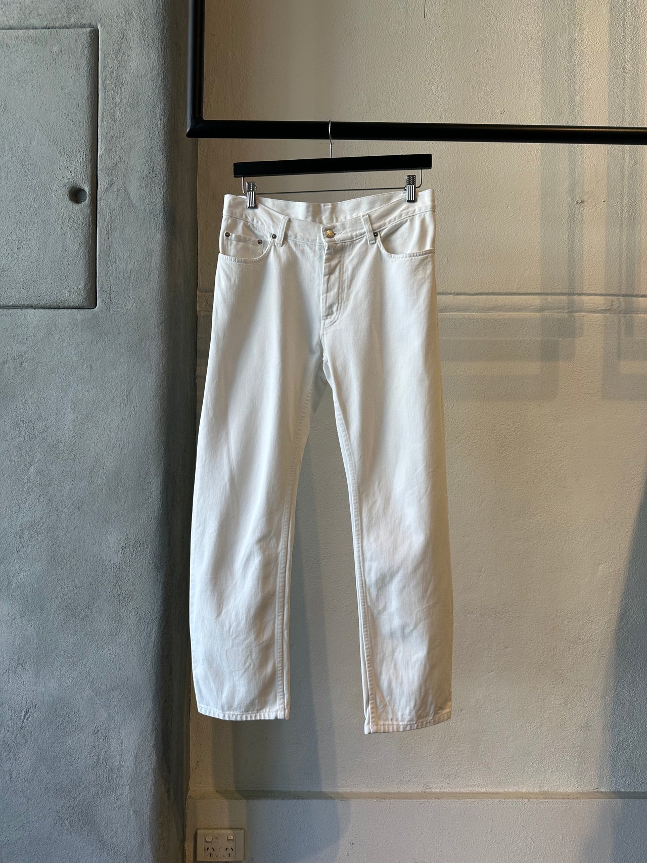 White Helmut Lang jeans hanging on a rack against a gray wall.
