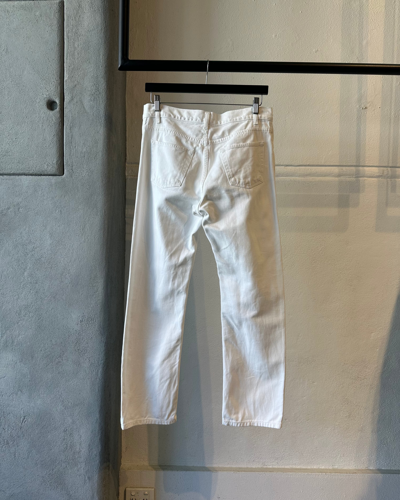 White Helmut Lang jeans hanging on a rack against a gray wall.
