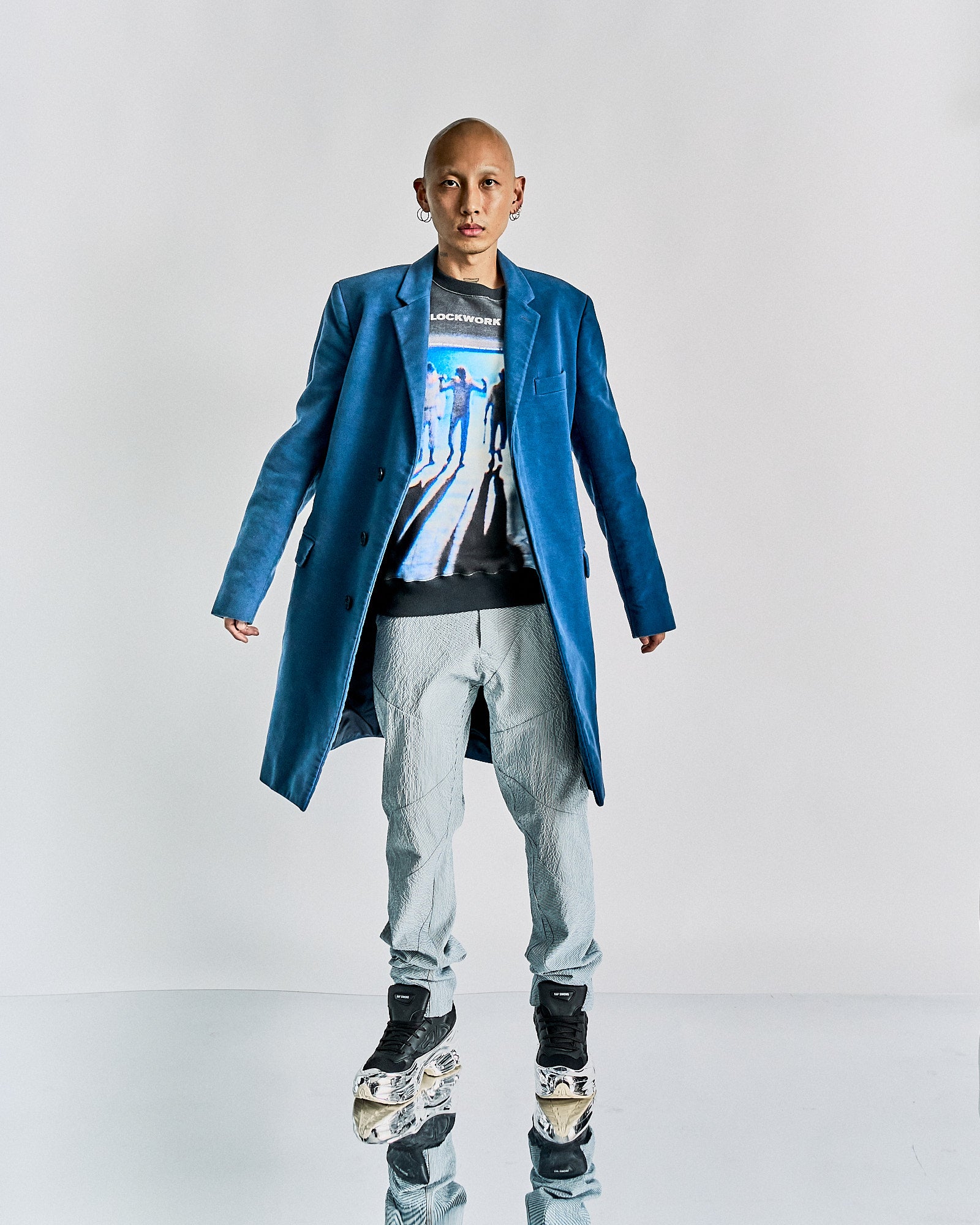 A person wearing a blue Helmut Lang SS 2005 knee-length coat with notch lapel, three-button closure, two hip pockets, one chest pocket, and four-button cuffs, standing on a reflective surface.