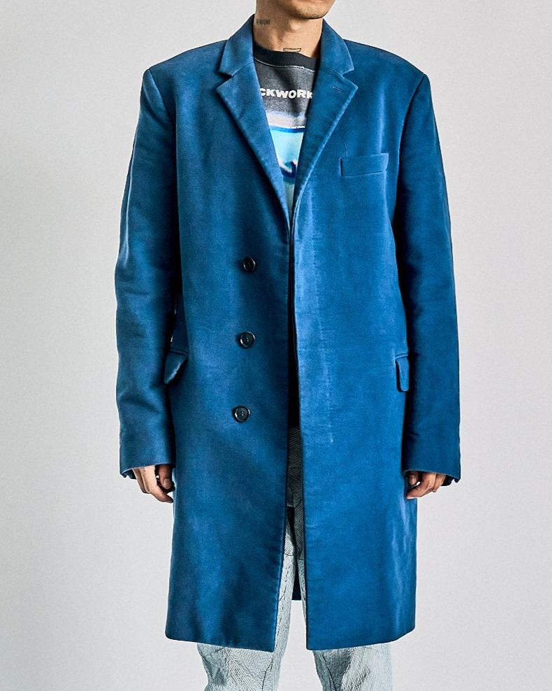 A person stands confidently against a plain background wearing a vibrant blue Helmut Lang coat, graphic t-shirt, textured pants, and unique platform sneakers.