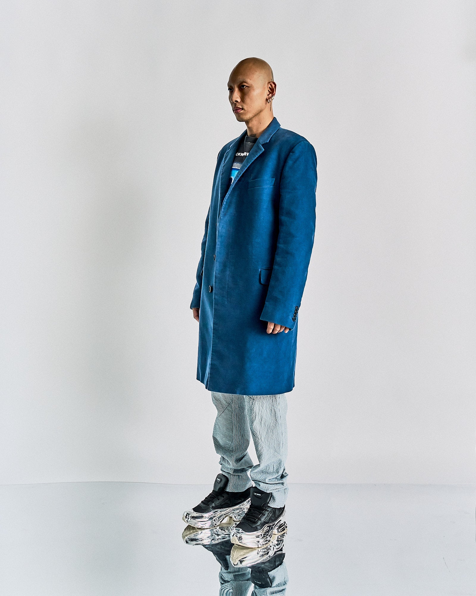 A person stands in a reflective space wearing a long, blue Helmut Lang coat, light gray pants, and unique metallic platform shoes, conveying a futuristic fashion style.